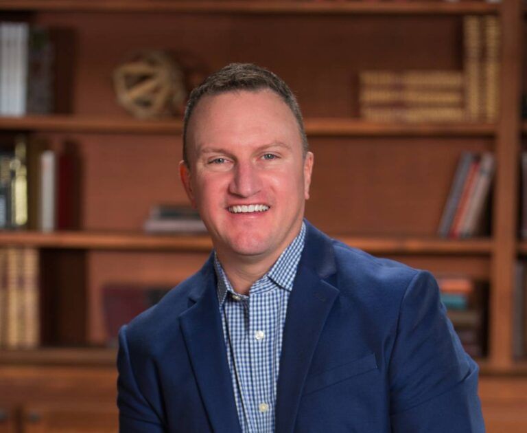 Man smiling, wearing a blue blazer over a checkered shirt, in front of a bookshelf.