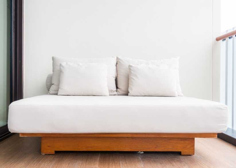 Un diván blanco con dos cojines cuadrados sobre una base de madera, apoyado contra una pared lisa.