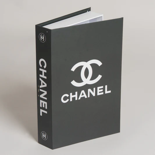 Una caja de almacenamiento negra con forma de libro, con el logotipo y el nombre de Chanel en blanco en la tapa y el lomo, sobre un fondo liso.