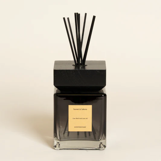 Un difusor de varillas negras en una botella de vidrio cuadrada con tapa negra y etiqueta dorada, sobre un fondo claro liso.