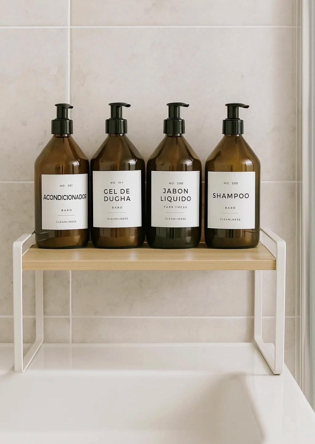 Cuatro frascos de color ámbar con dosificador, etiquetados con artículos de tocador, reposan sobre una repisa de madera clara y metal blanco, apoyada contra una pared alicatada del baño.