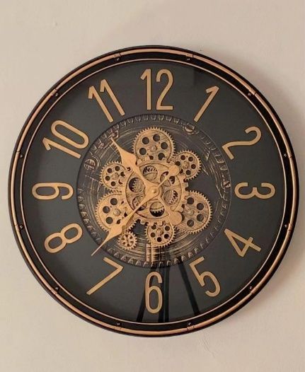 Un reloj de pared redondo y negro con números dorados y engranajes decorativos a la vista en el centro.