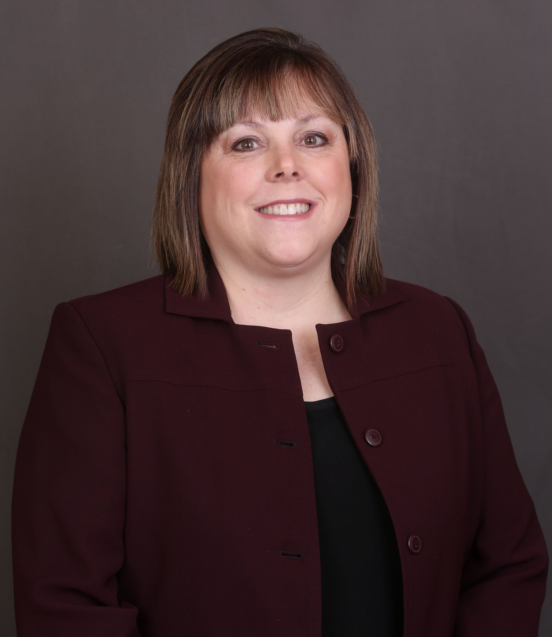 Becky Dodson Woman smiling in a dark red blazer and black top against a grey background.