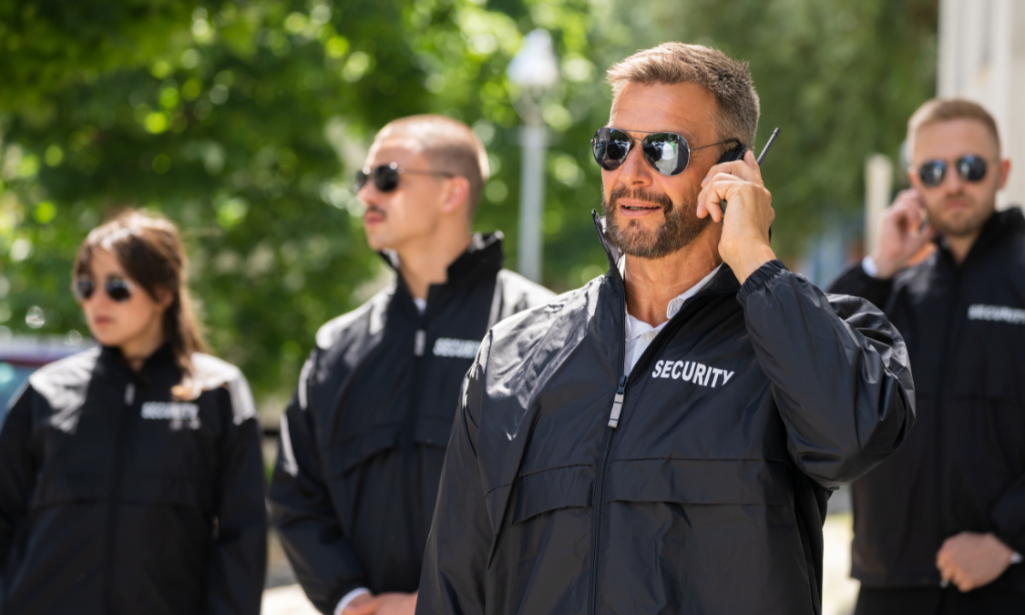 Security personnel in black jackets and sunglasses, one talking on a radio, outside.