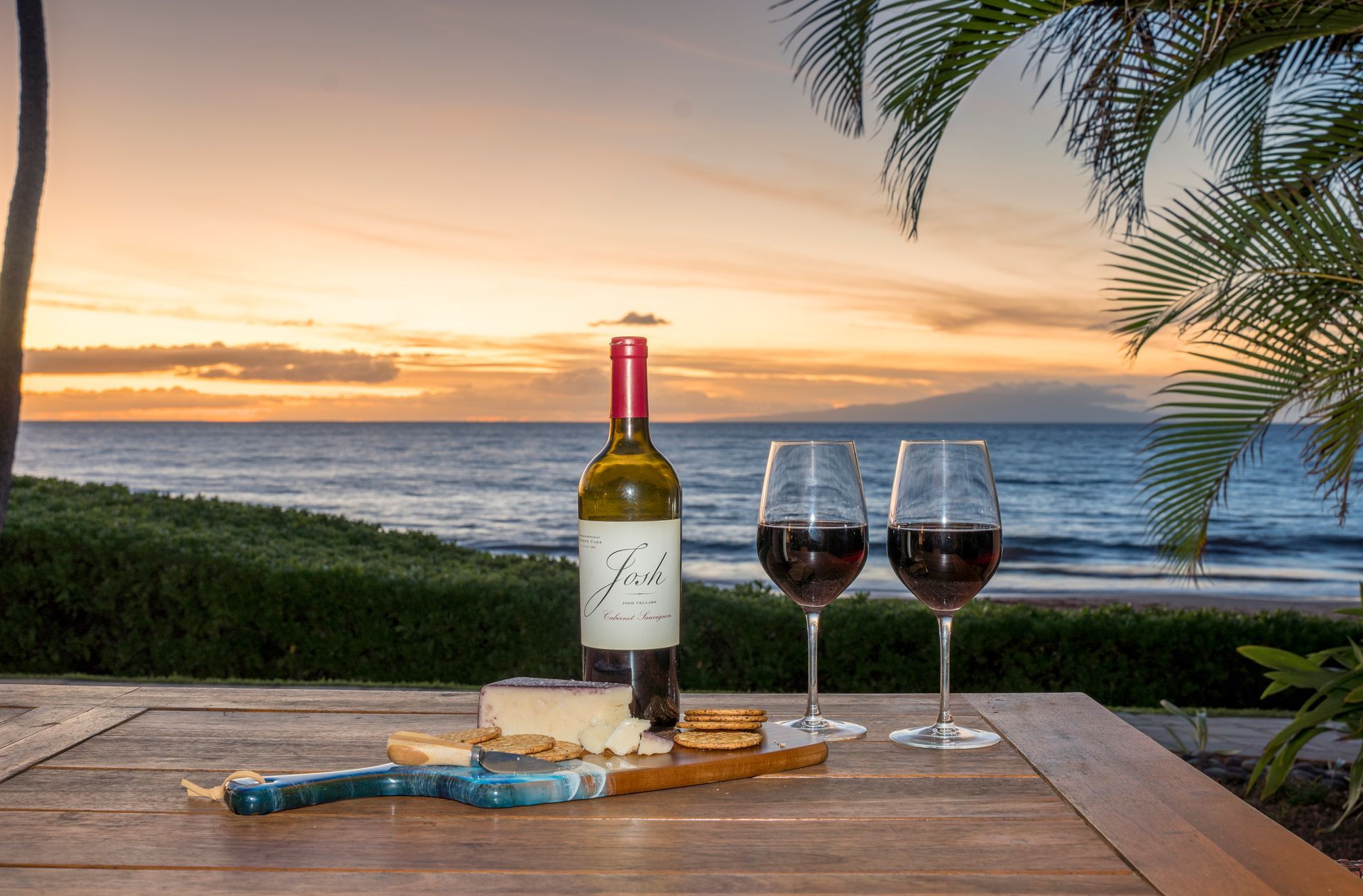 Wine and cheese on a table with ocean sunset background; two filled glasses, bottle, and a cutting board.