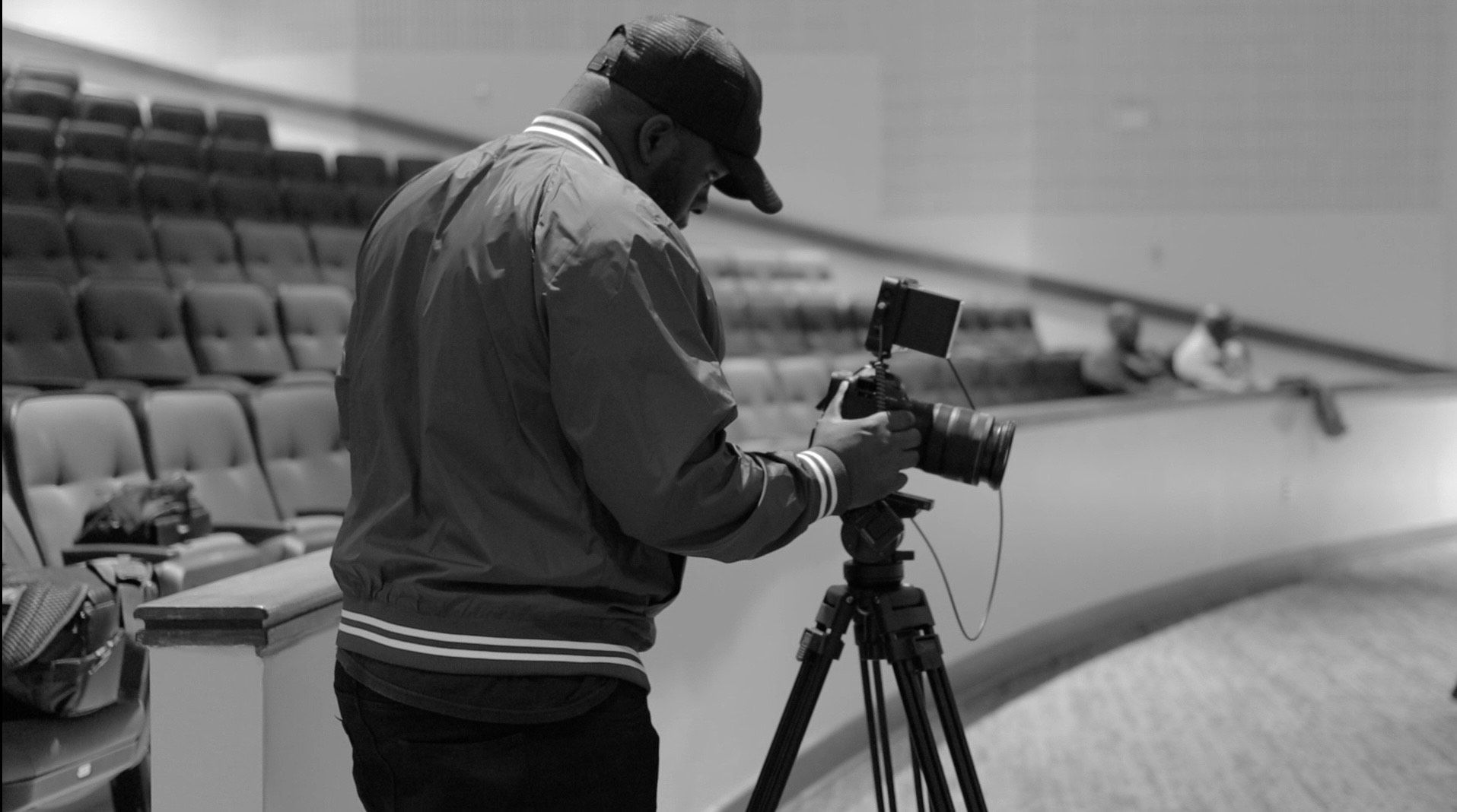 A person holding a camera, outdoors near a building with foliage.
