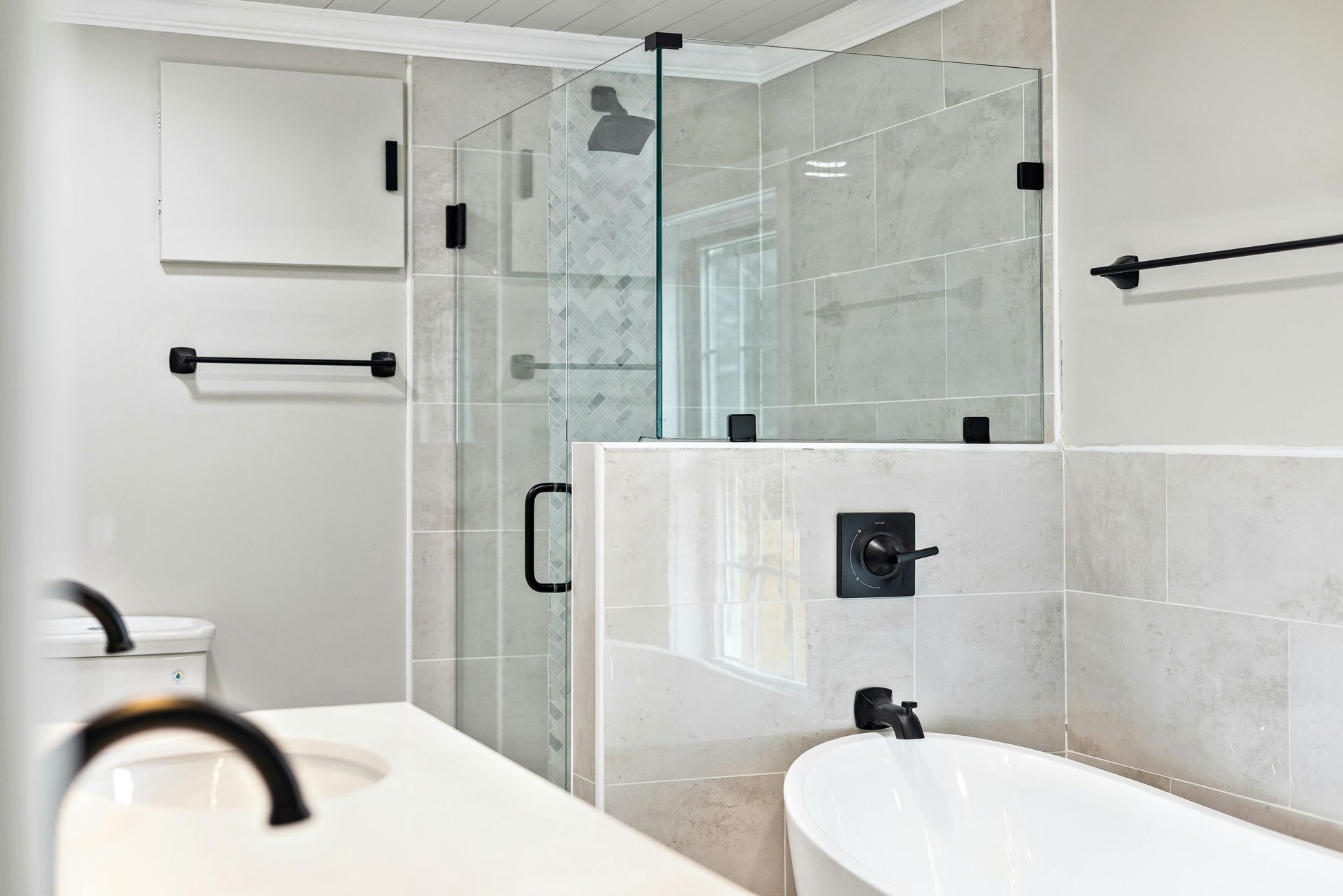 Modern bathroom with a glass shower, white tub, and light-colored tile. Black fixtures and hardware.
