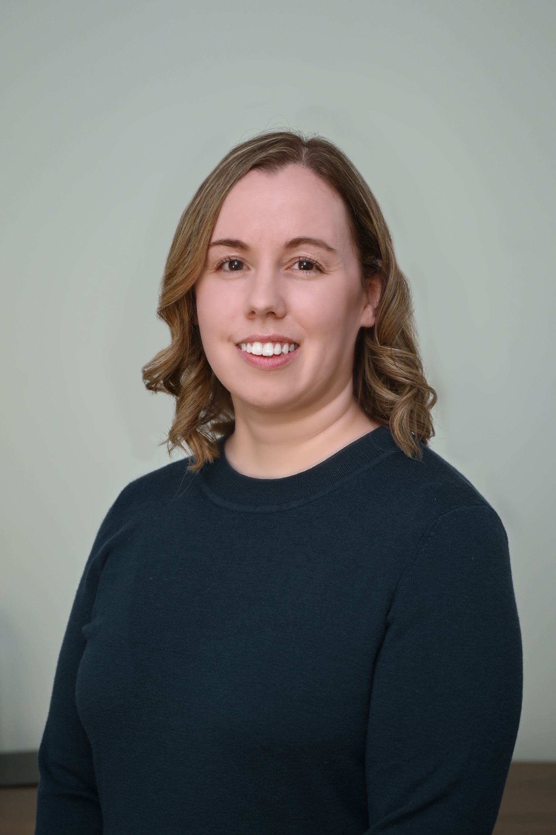 Woman with shoulder-length, wavy blonde hair, smiling, wearing a dark teal sweater, against a light gray background.
