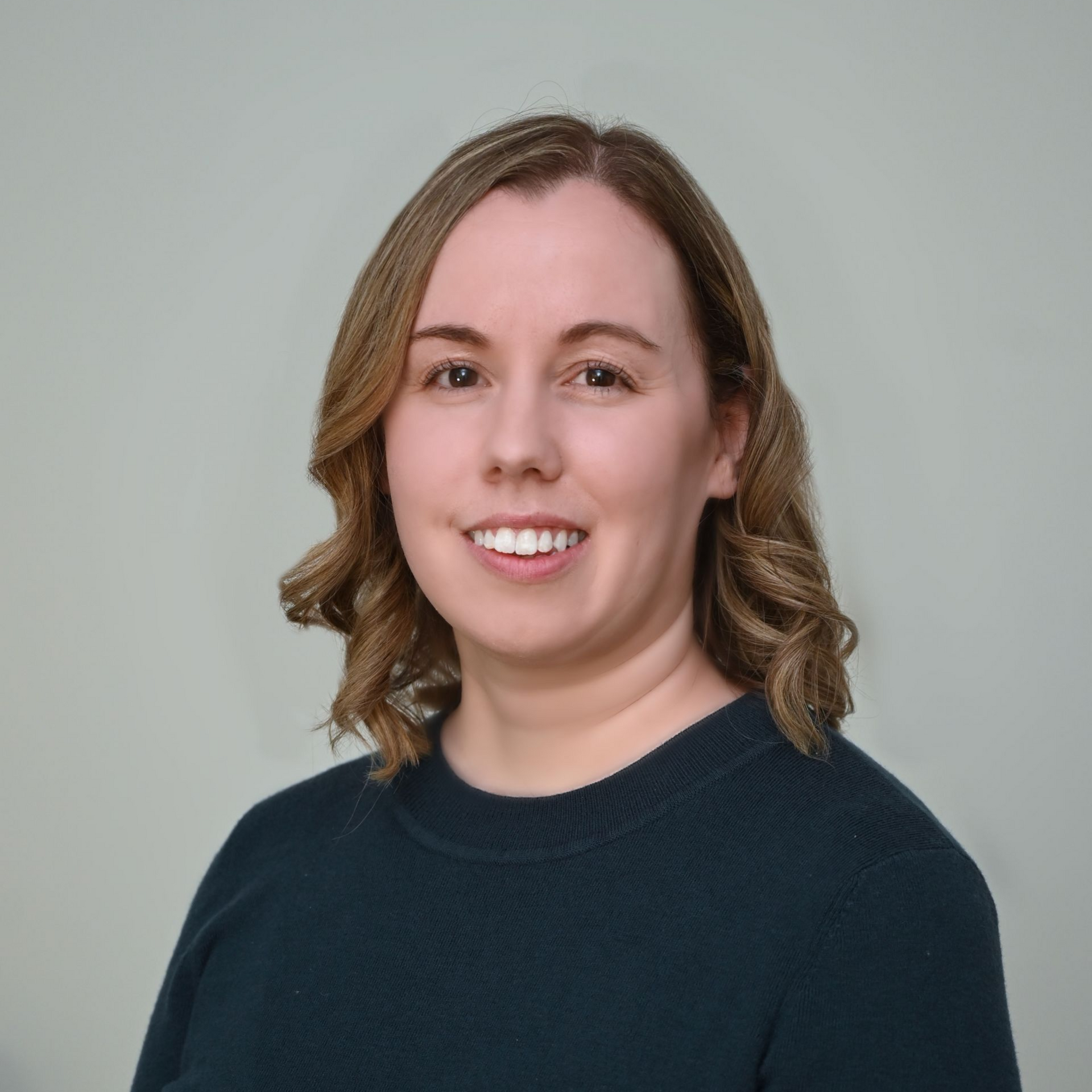 Woman with shoulder-length, wavy blonde hair, smiling, wearing a dark teal sweater, against a light gray background.