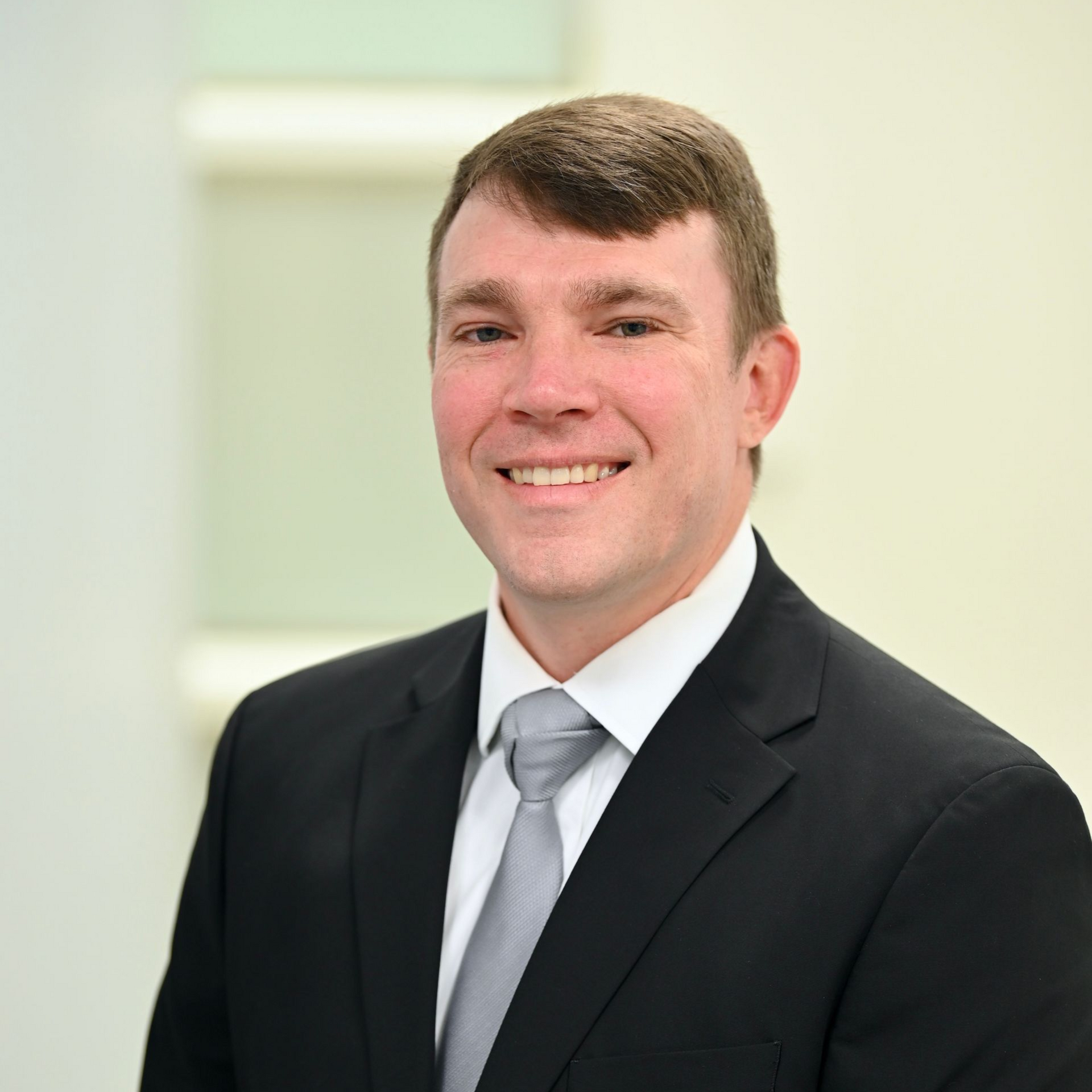 Man in black suit and tie, smiling, in a bright office setting.
