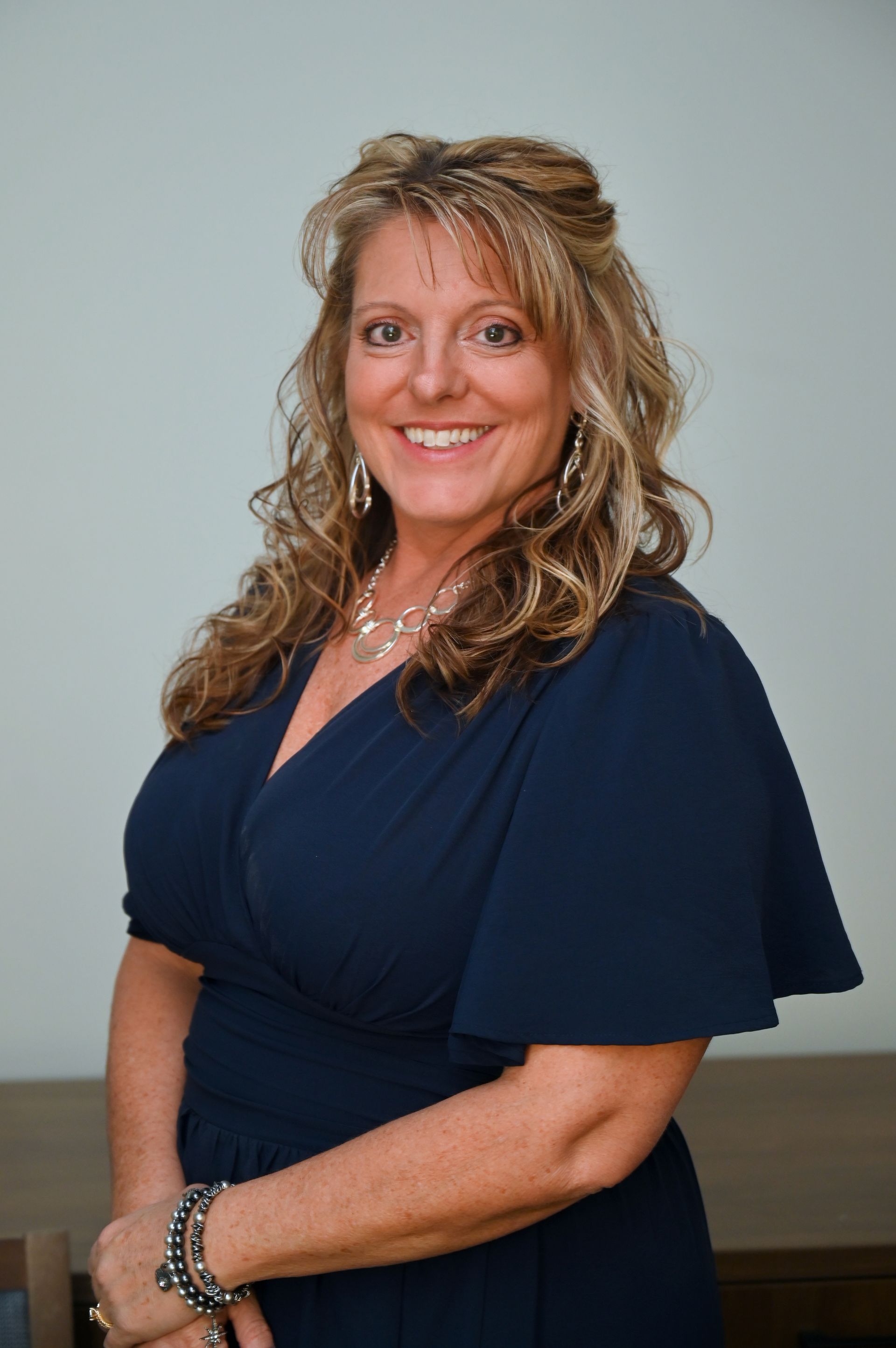 Woman in a navy dress smiles, looking at the camera. She has long curly hair, and wears jewelry.