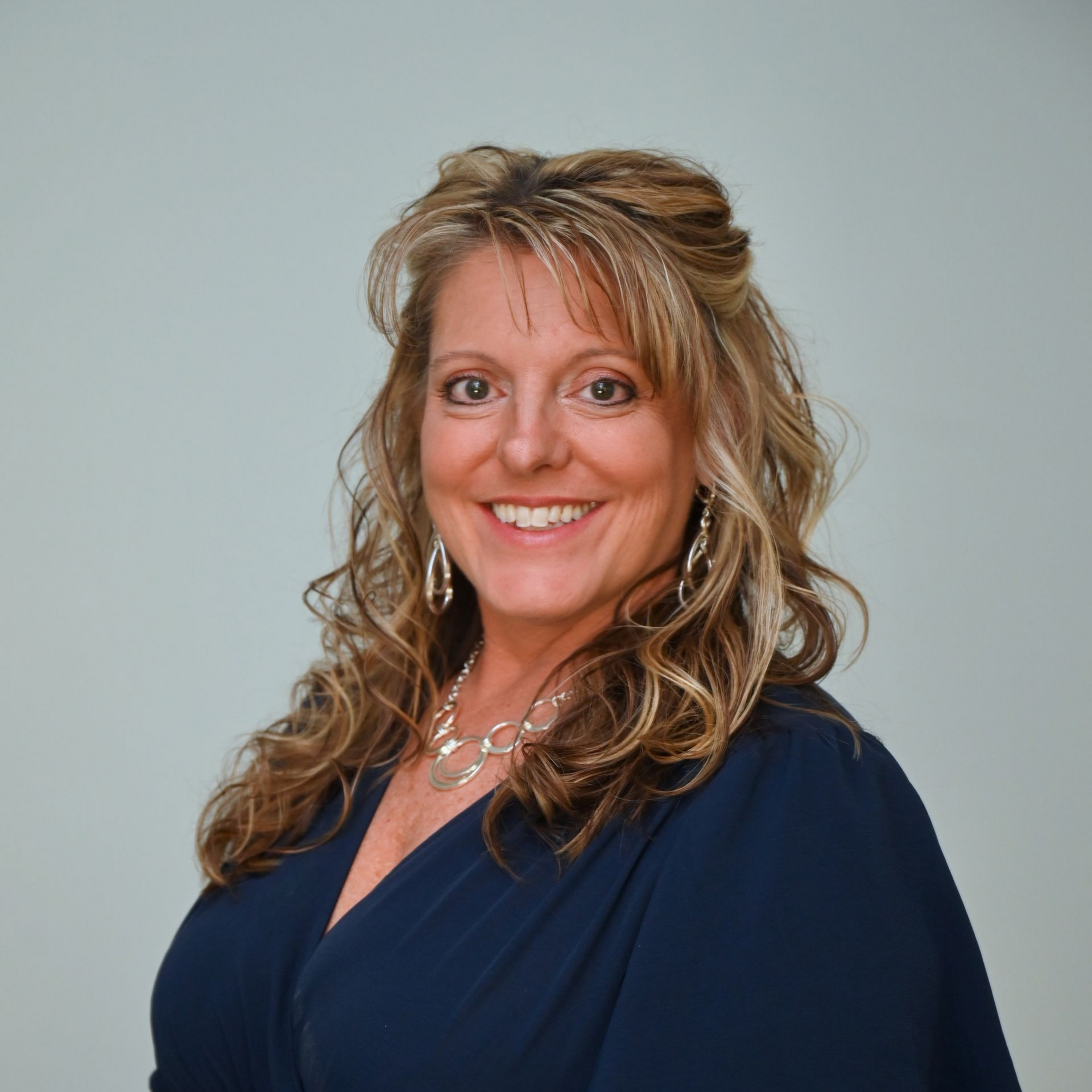 Woman in a navy dress smiles, looking at the camera. She has long curly hair, and wears jewelry.