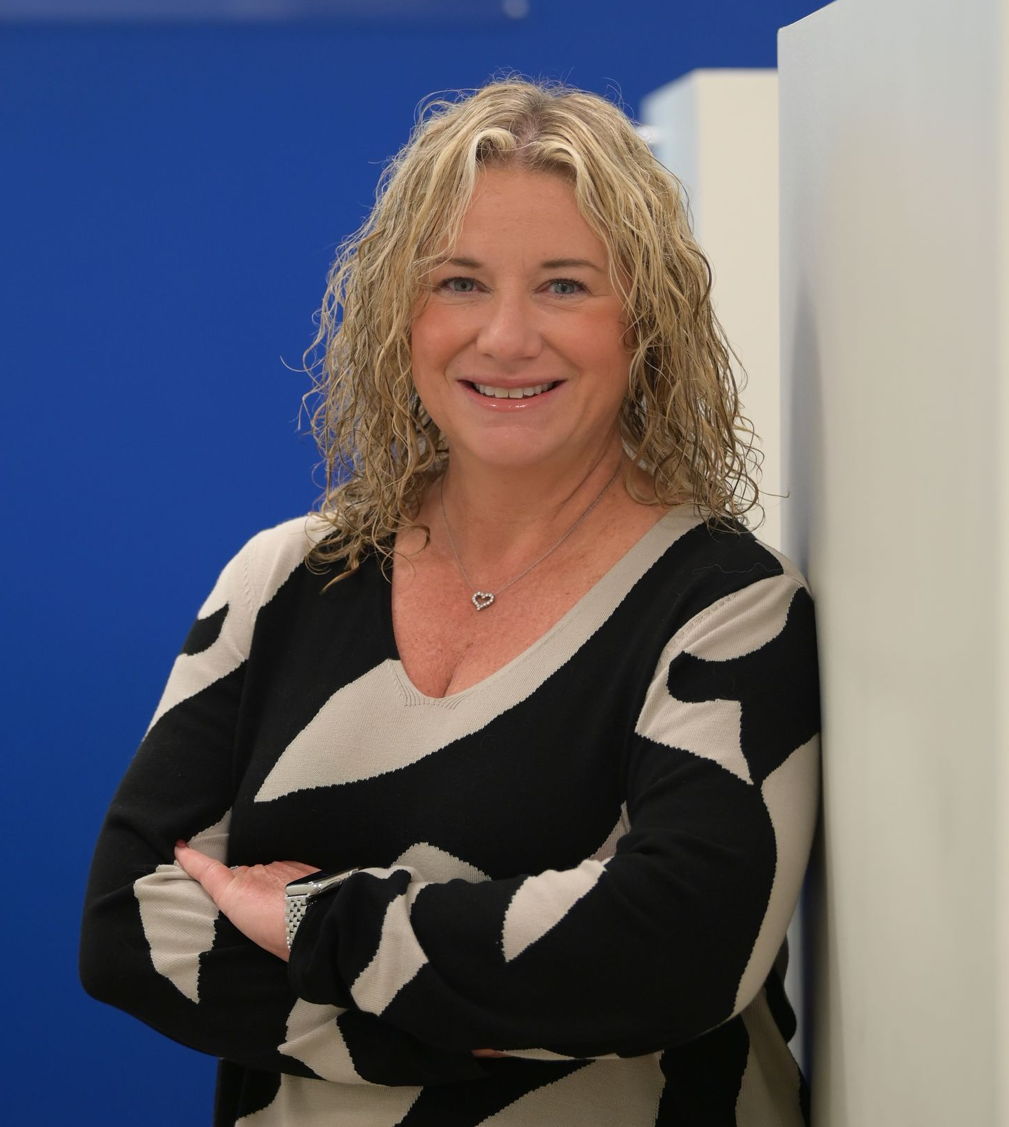Woman in black and white patterned top with arms crossed, in office setting with a blue wall and logo.