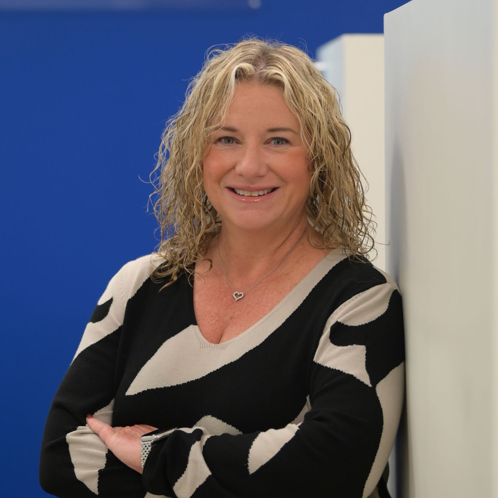 Woman in black and white patterned top with arms crossed, in office setting with a blue wall and logo.