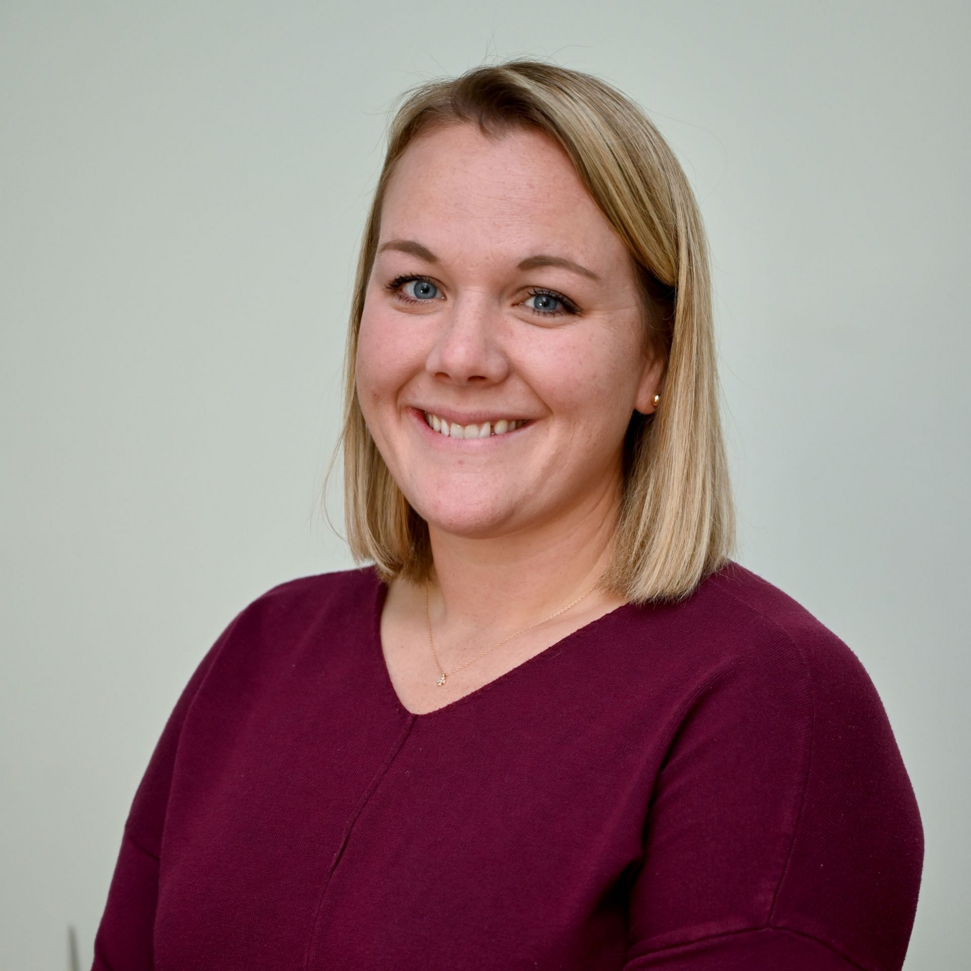 Woman with short blonde hair smiles, wearing a burgundy v-neck sweater, in front of a light wall.