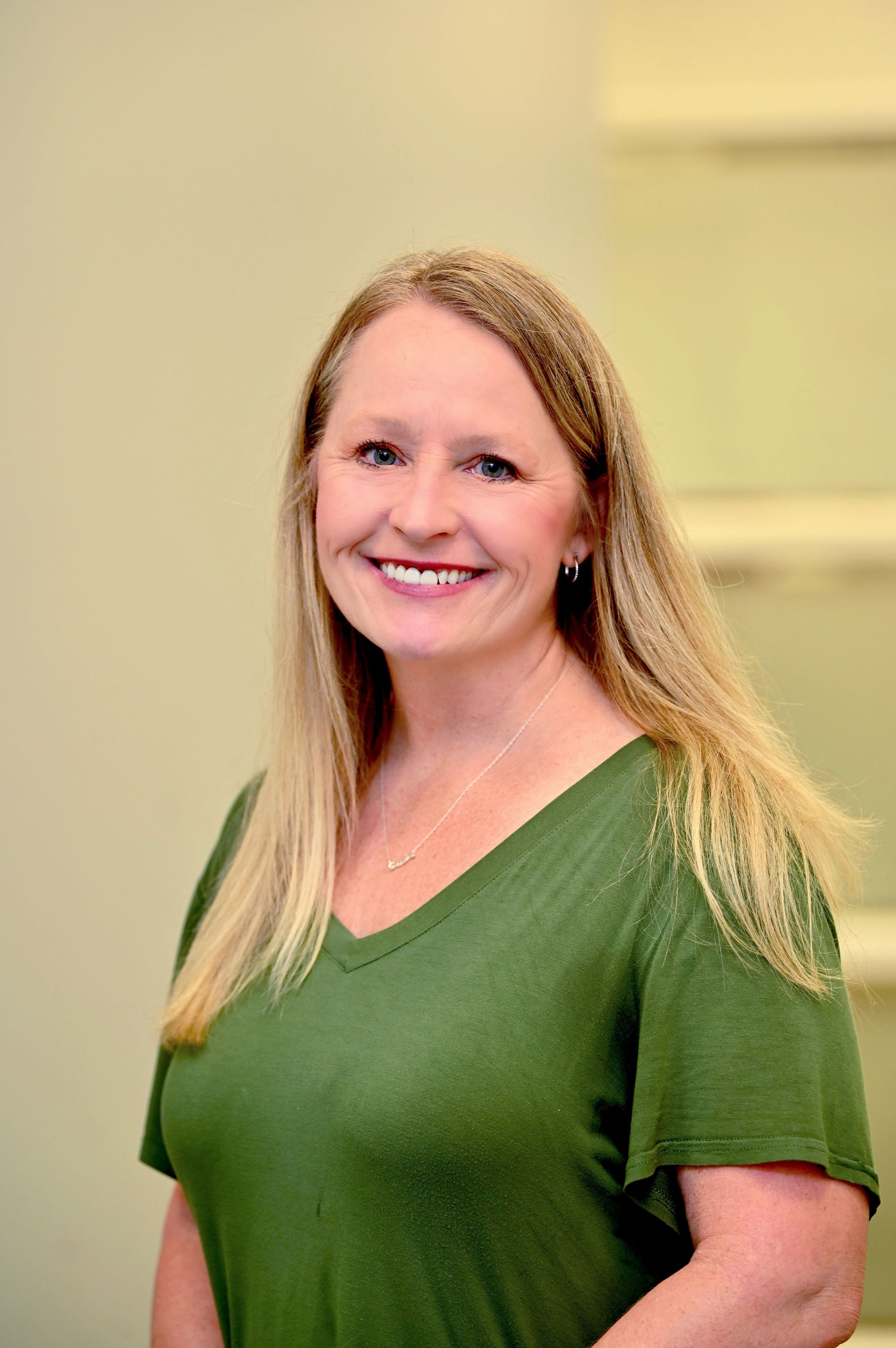 Woman with blonde hair wearing a green v-neck shirt smiles in a neutral-toned room.