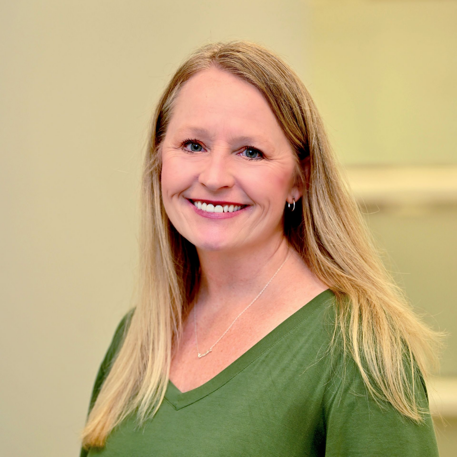 Woman with blonde hair wearing a green v-neck shirt smiles in a neutral-toned room.