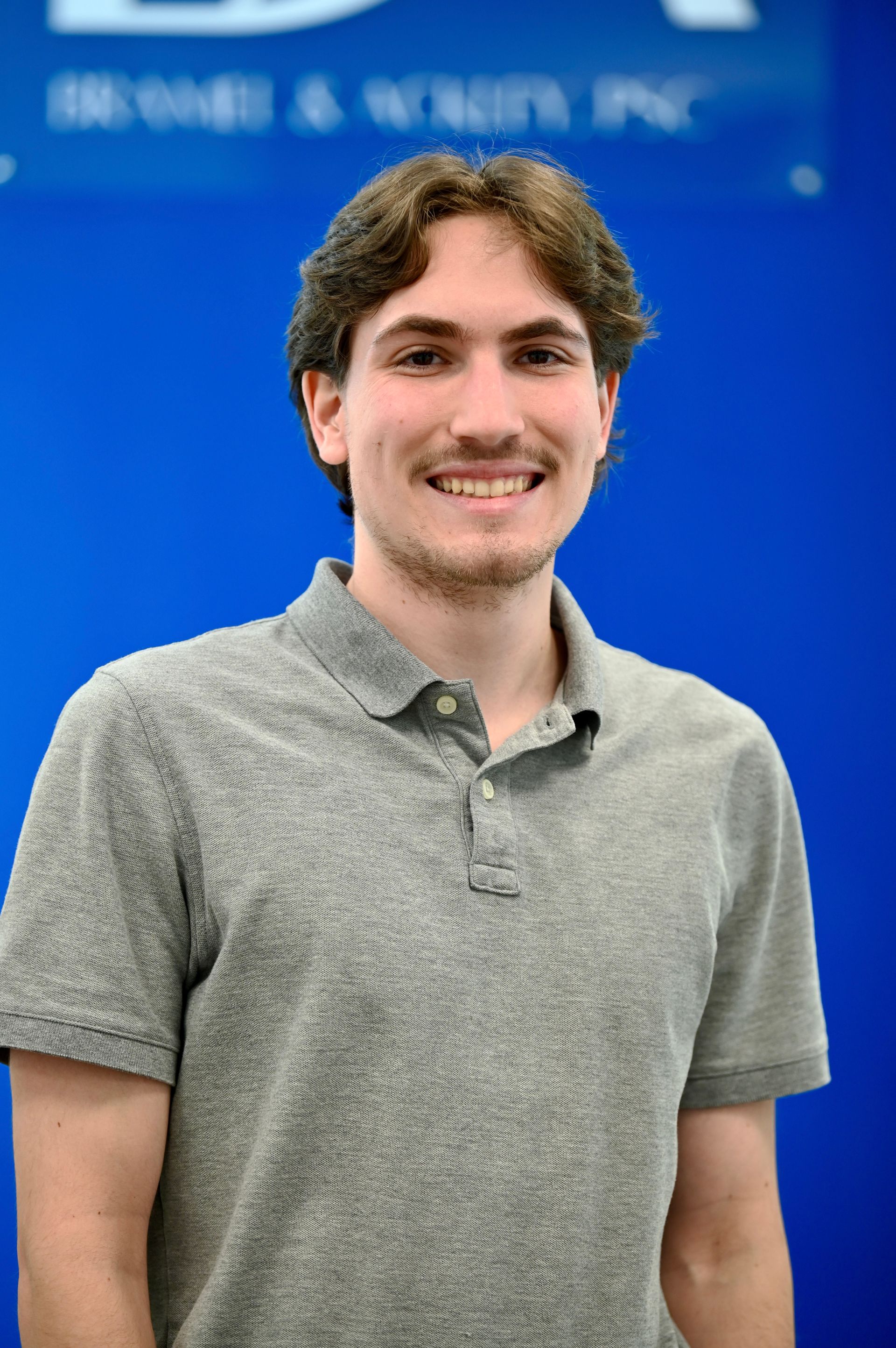 Man smiling, wearing a gray polo shirt, in front of a blue background with text.