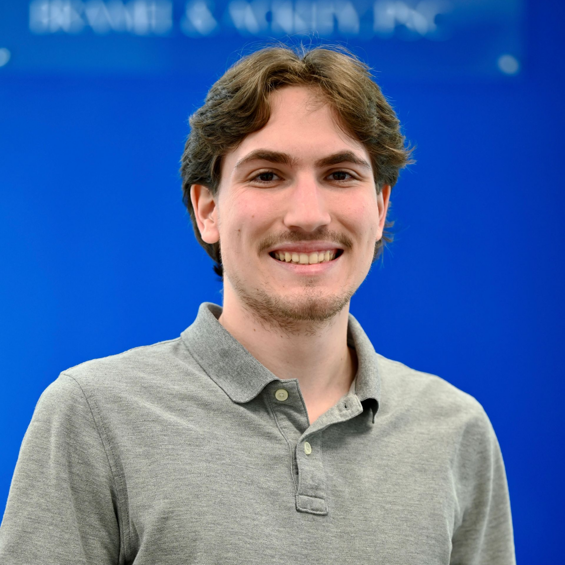 Man smiling, wearing a gray polo shirt, in front of a blue background with text.