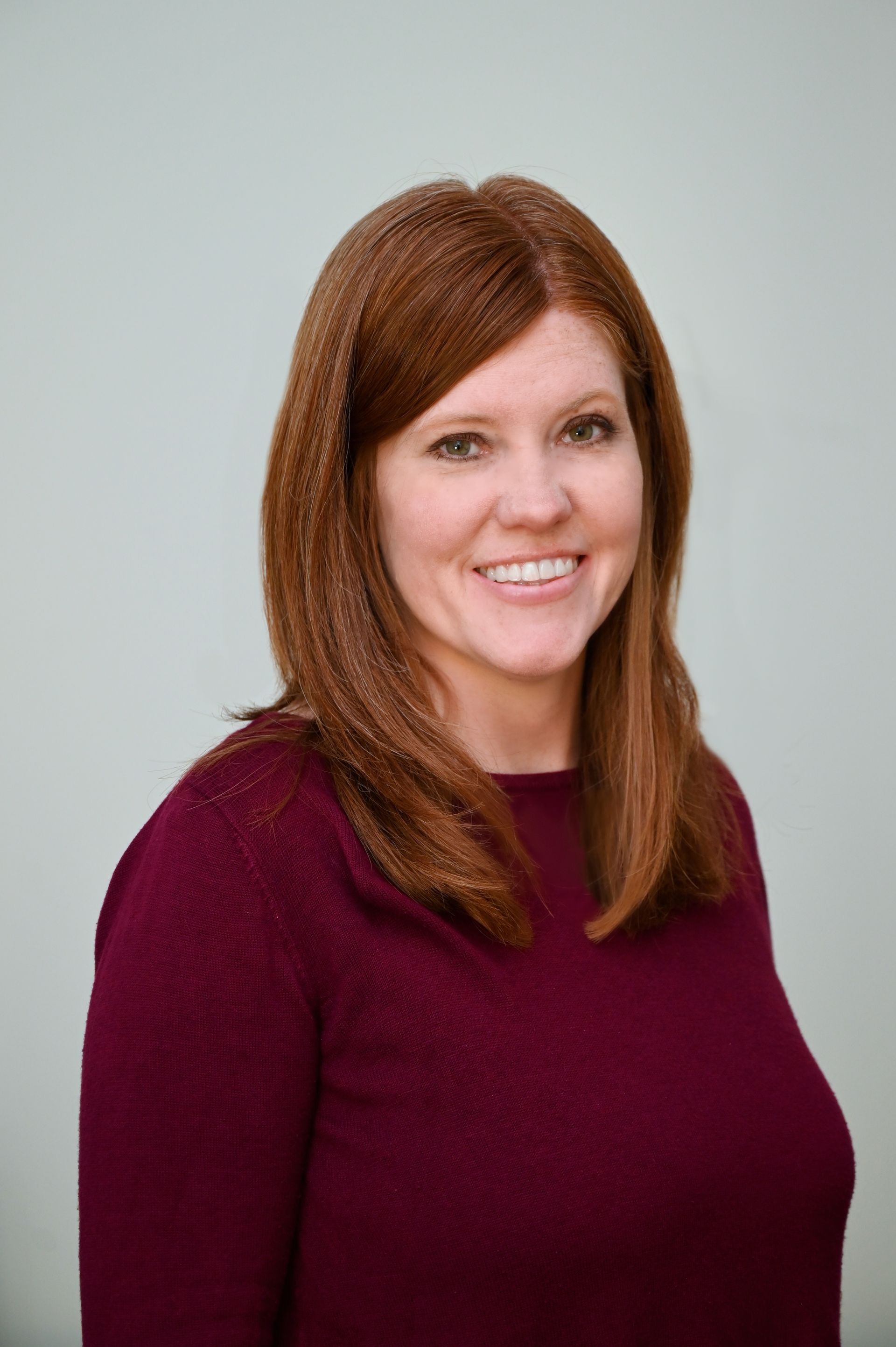 Woman with red hair smiling, wearing a maroon sweater, standing against a light blue background.