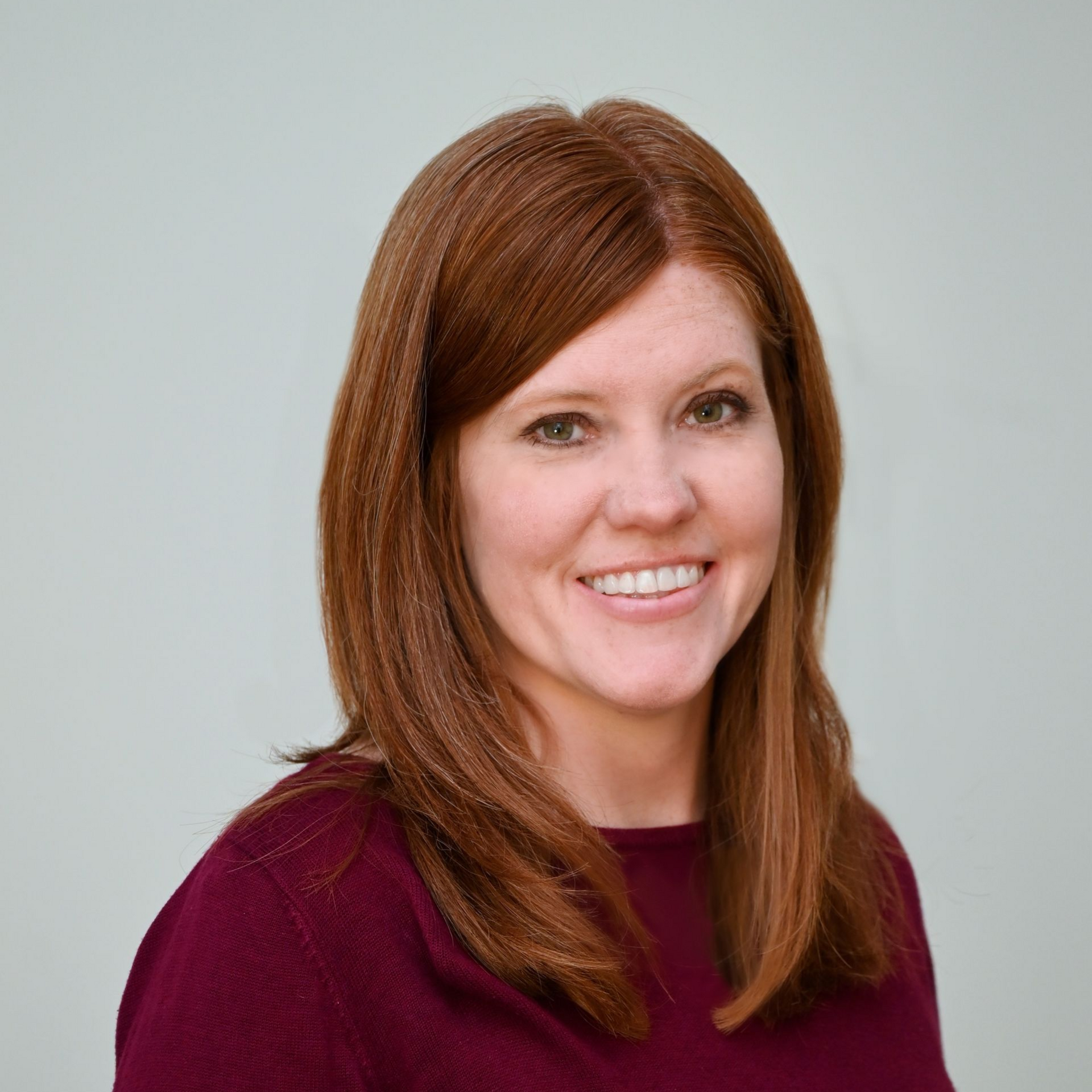 Woman with red hair smiling, wearing a maroon sweater, standing against a light blue background.