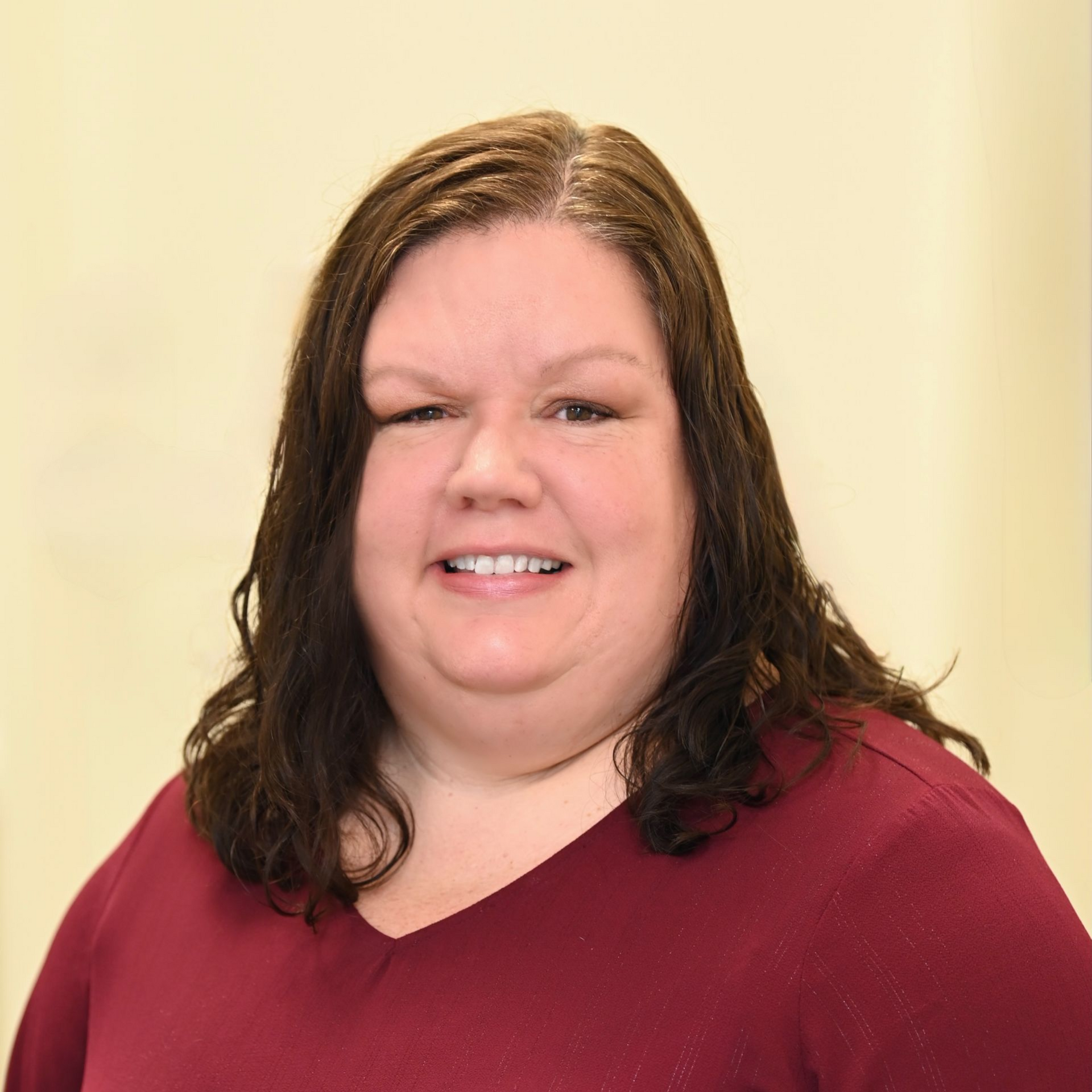 Woman with dark hair smiles, wearing a burgundy v-neck sweater, in front of a plain background.