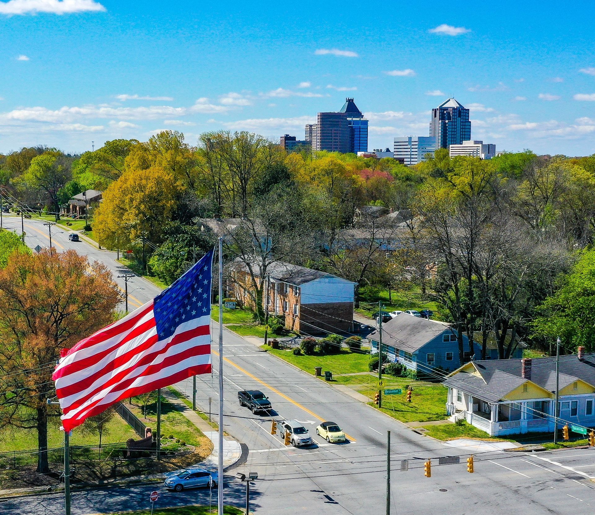 American flag waving over a city street lined with buildings and trees, sunny day.