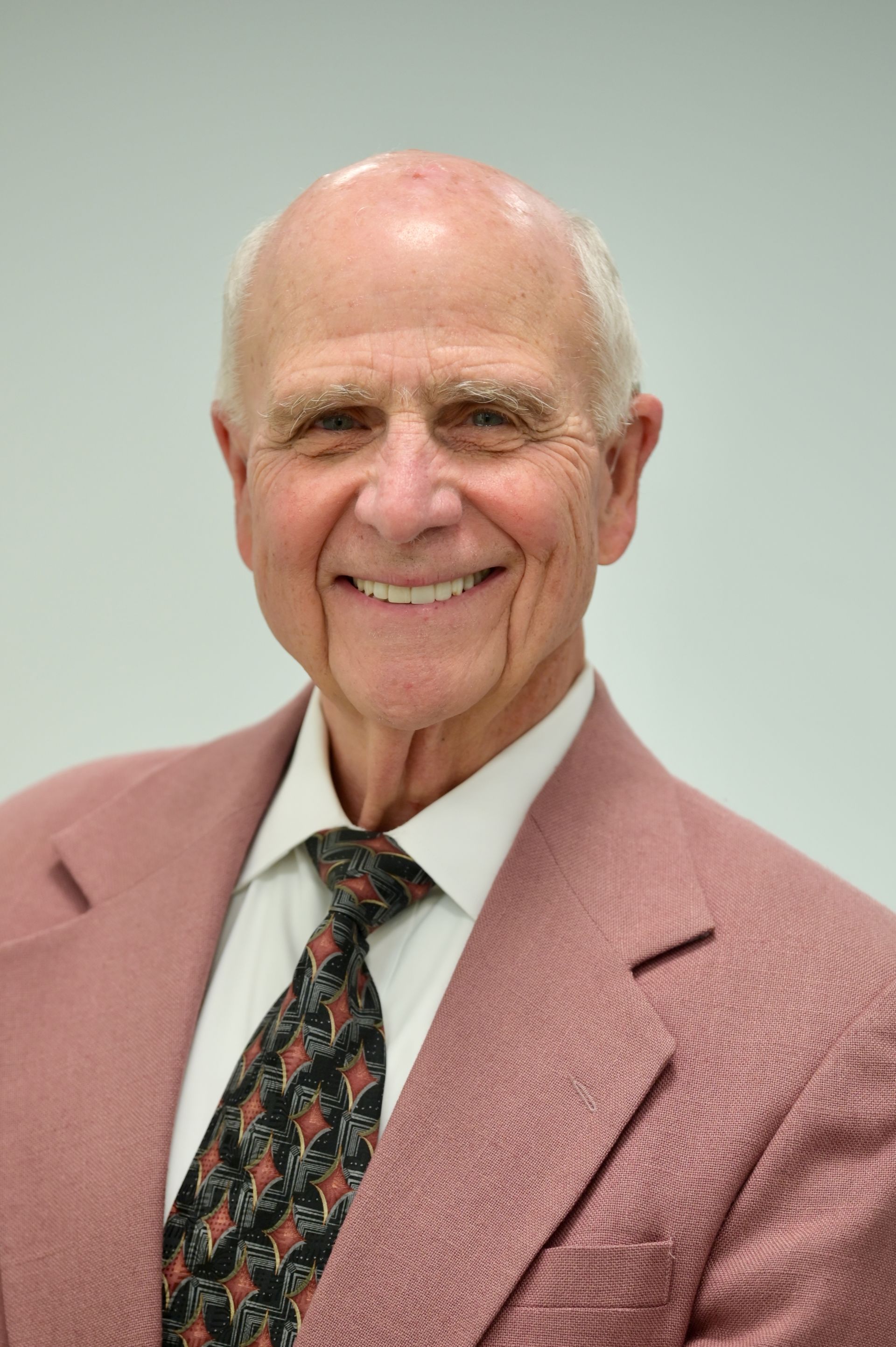 Smiling older man in a pink blazer and patterned tie, against a white background.