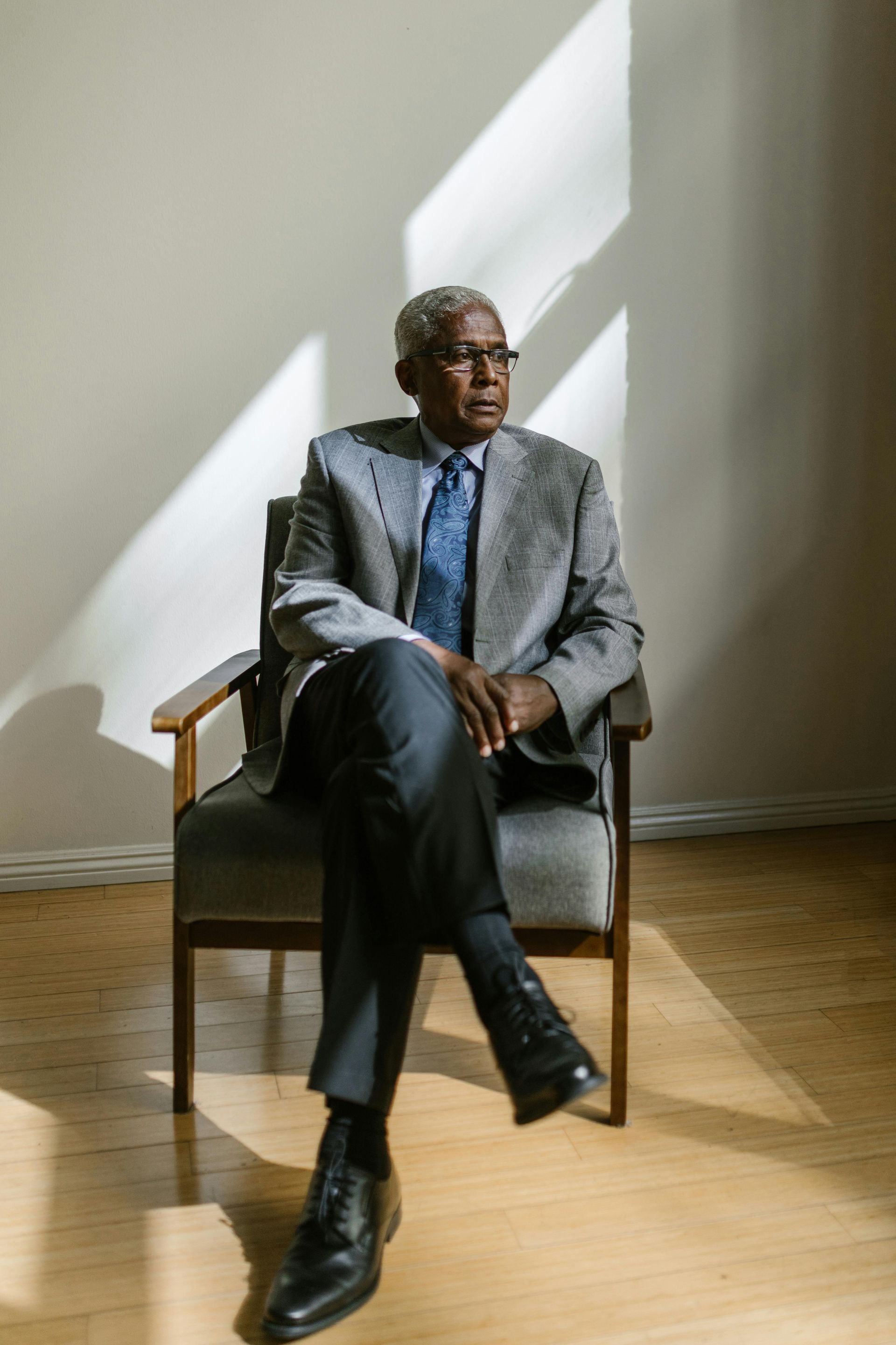 Man in a gray suit sits in a chair, looking away; sunlight casts stripes on the wall behind him.