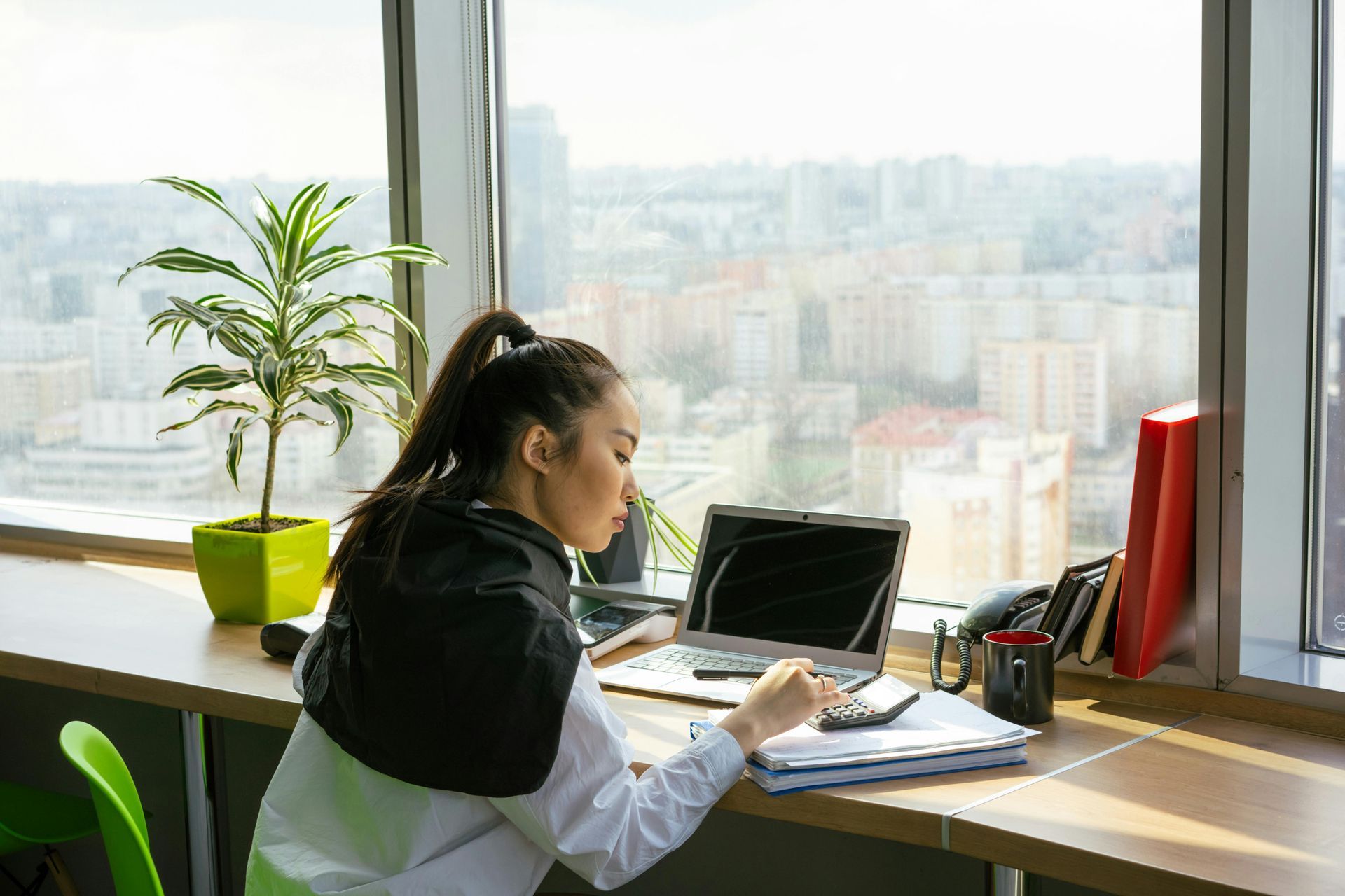 Woman working at a desk near a window with city view, using a laptop, calculator, and documents.