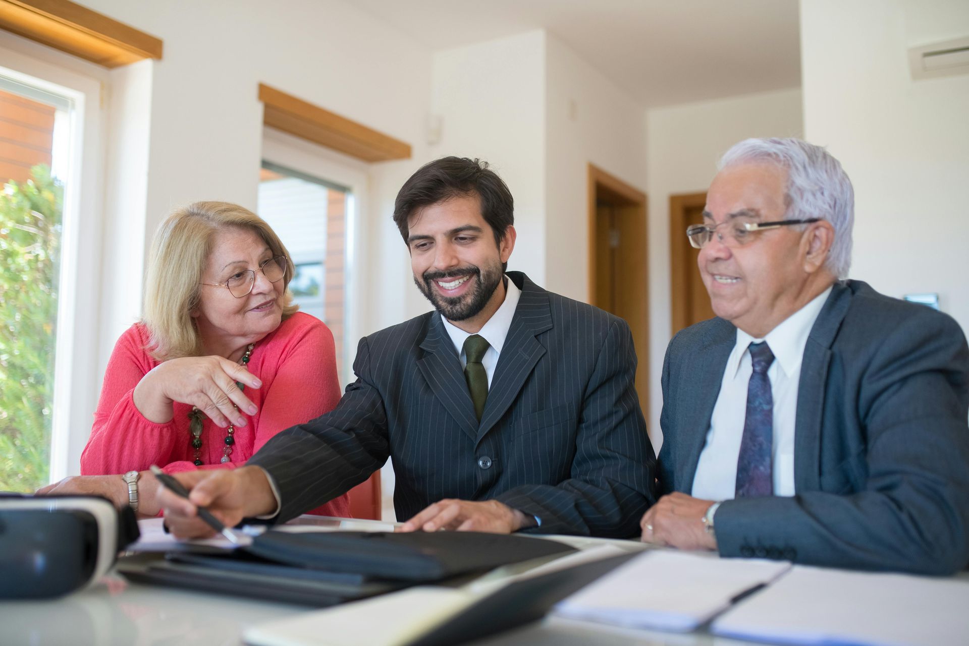 A man in a suit points to papers as an elderly couple watches; a bright room with window.