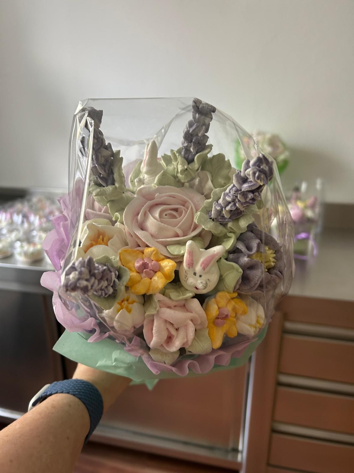 Eine Hand hält eine durchsichtige Geschenktüte mit pastellfarbenen Kunstblumen und Lavendelakzenten.