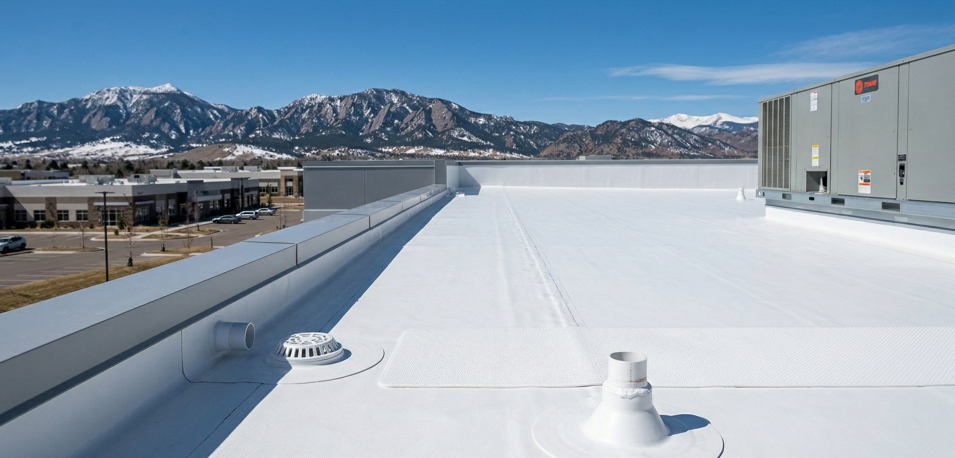 A view from a flat, white rooftop featuring HVAC equipment, a drainage vent, and distant snow-capped mountains.
