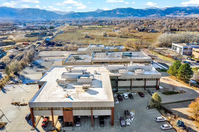 An aerial view of a multi-story office building complex with parking lots, surrounding fields, and mountains in the back.