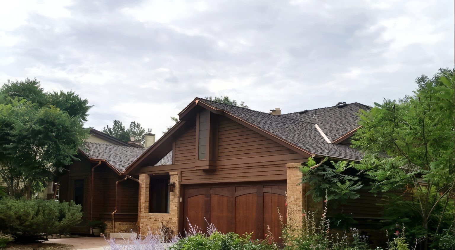 A single-story home with a tan stone facade, dark wood garage doors, and shingled roof, nestled among trees.