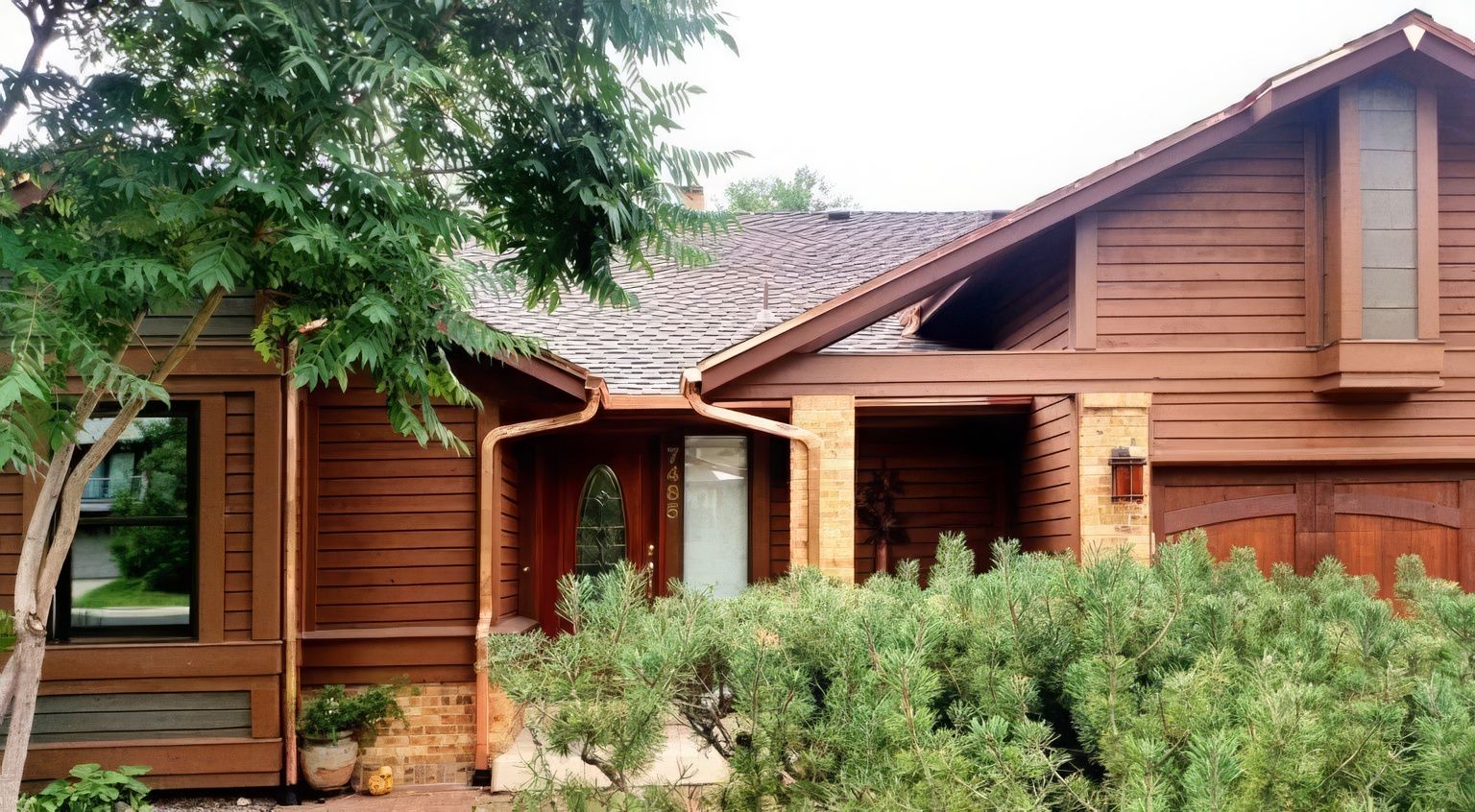 A single-story brown wooden house with a shingled roof, stone accents, and a large front garden with a tree.