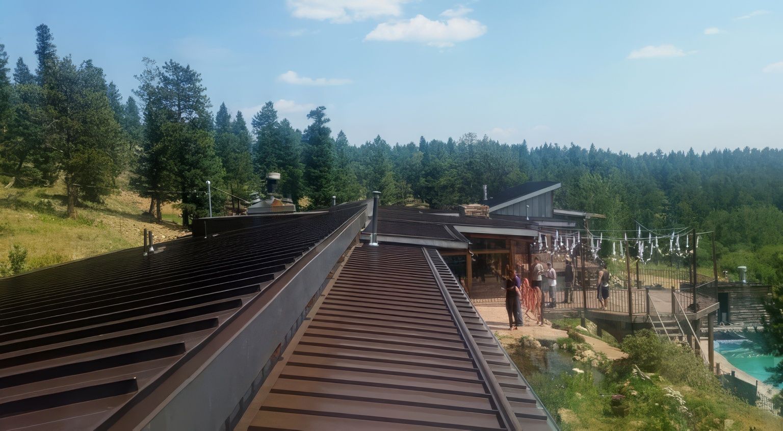 A metal roof overlooks an outdoor terrace and a swimming pool nestled in a forested, hilly landscape under a blue sky.