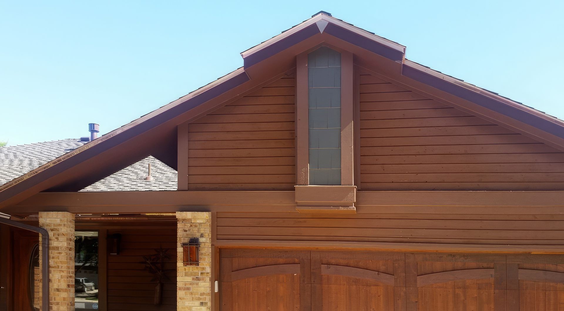 A brown house facade with a stone pillar, horizontal wood siding, a tall narrow window, and a wooden garage door.