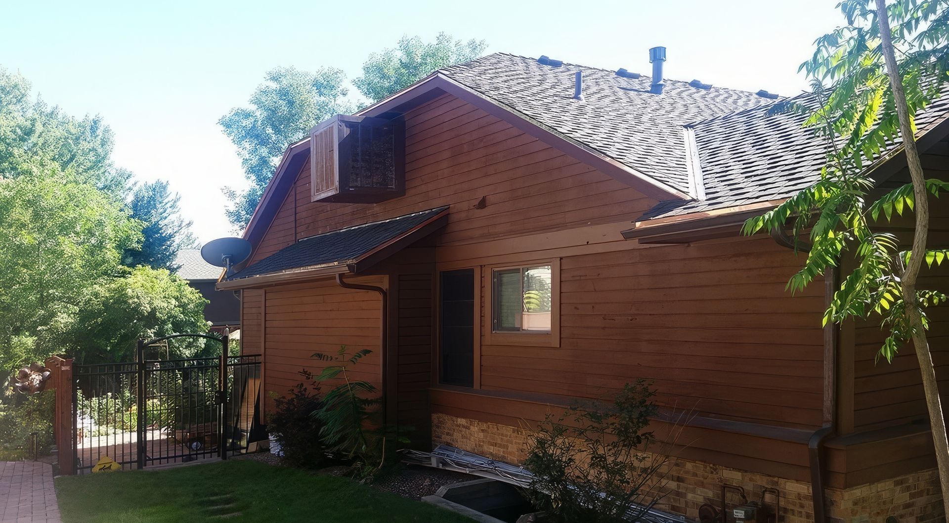 A brown house with horizontal siding, a stone base, a roof with dark shingles, and a window, surrounded by trees and grass.