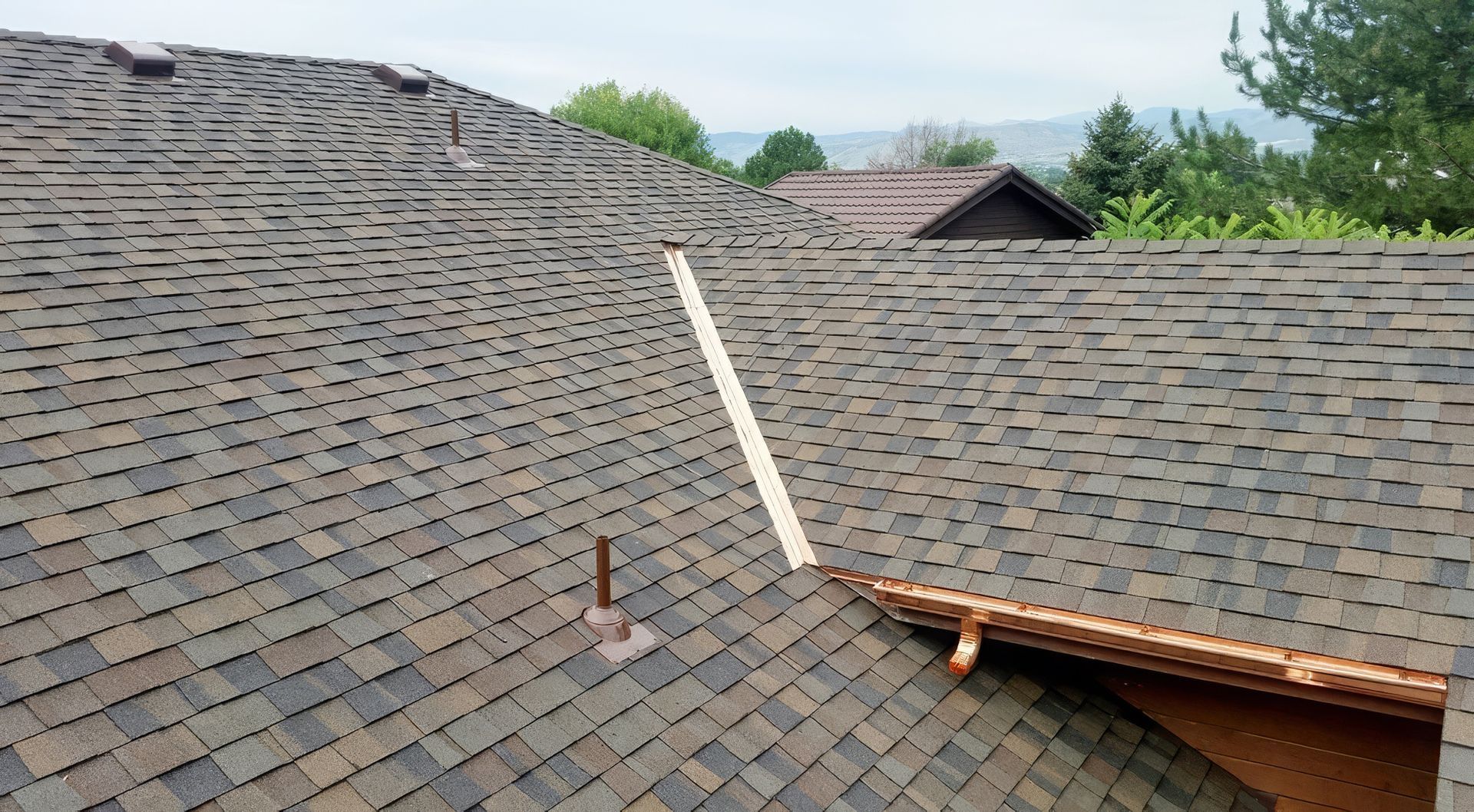An asphalt shingle roof featuring a light-colored valley flashing and an exposed pipe vent against a suburban background.