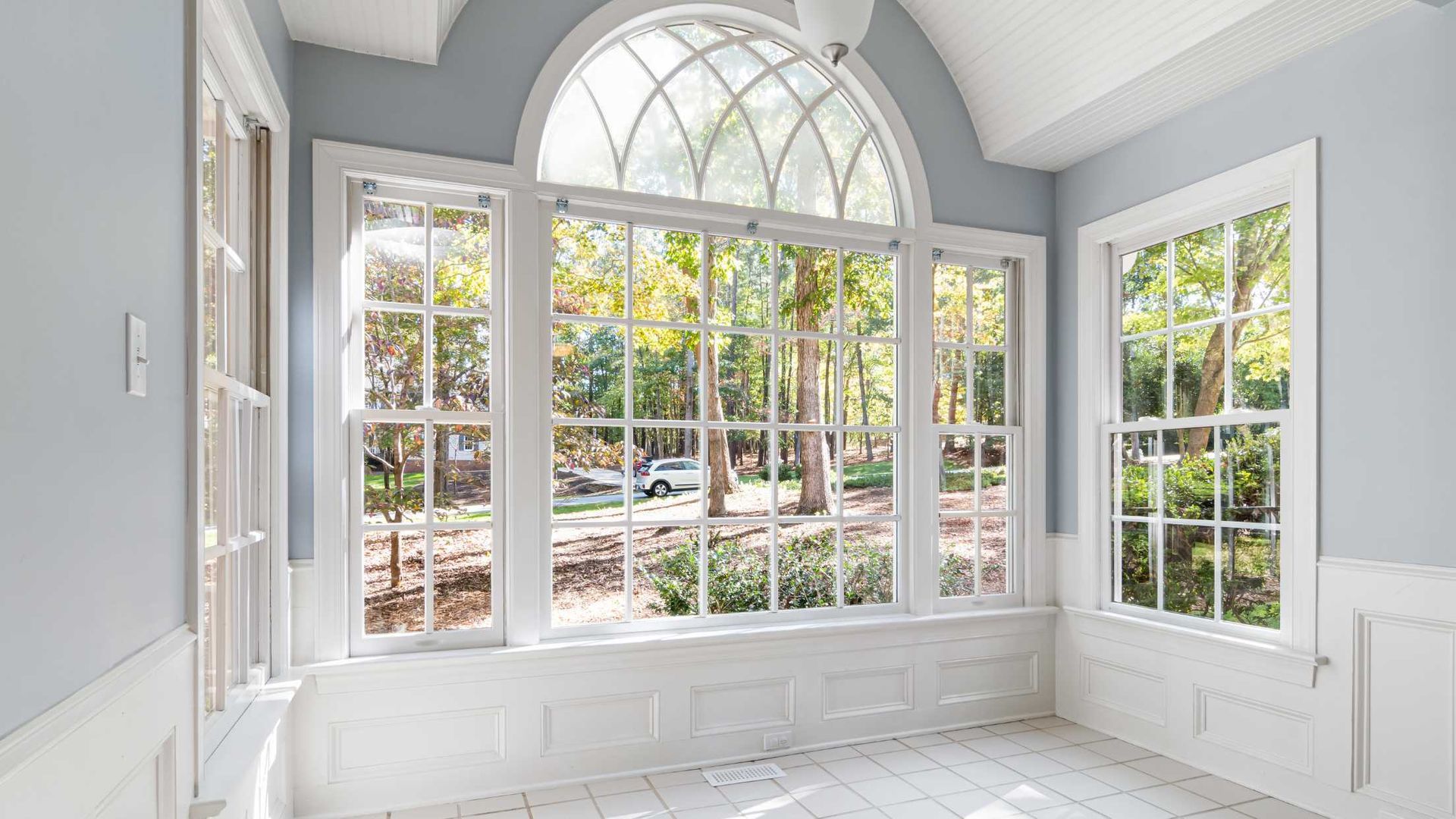 A bright room with light blue walls and white wainscoting features a large arched window looking out at trees.