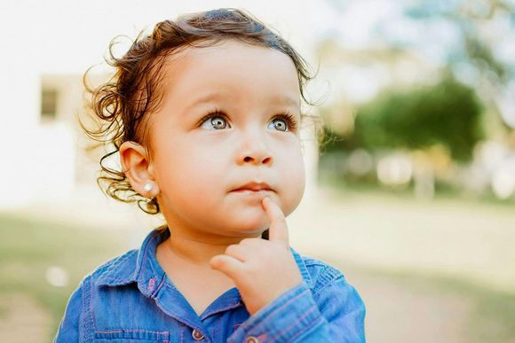 Child with curly hair and blue eyes, looking up with finger on chin, outdoors.
