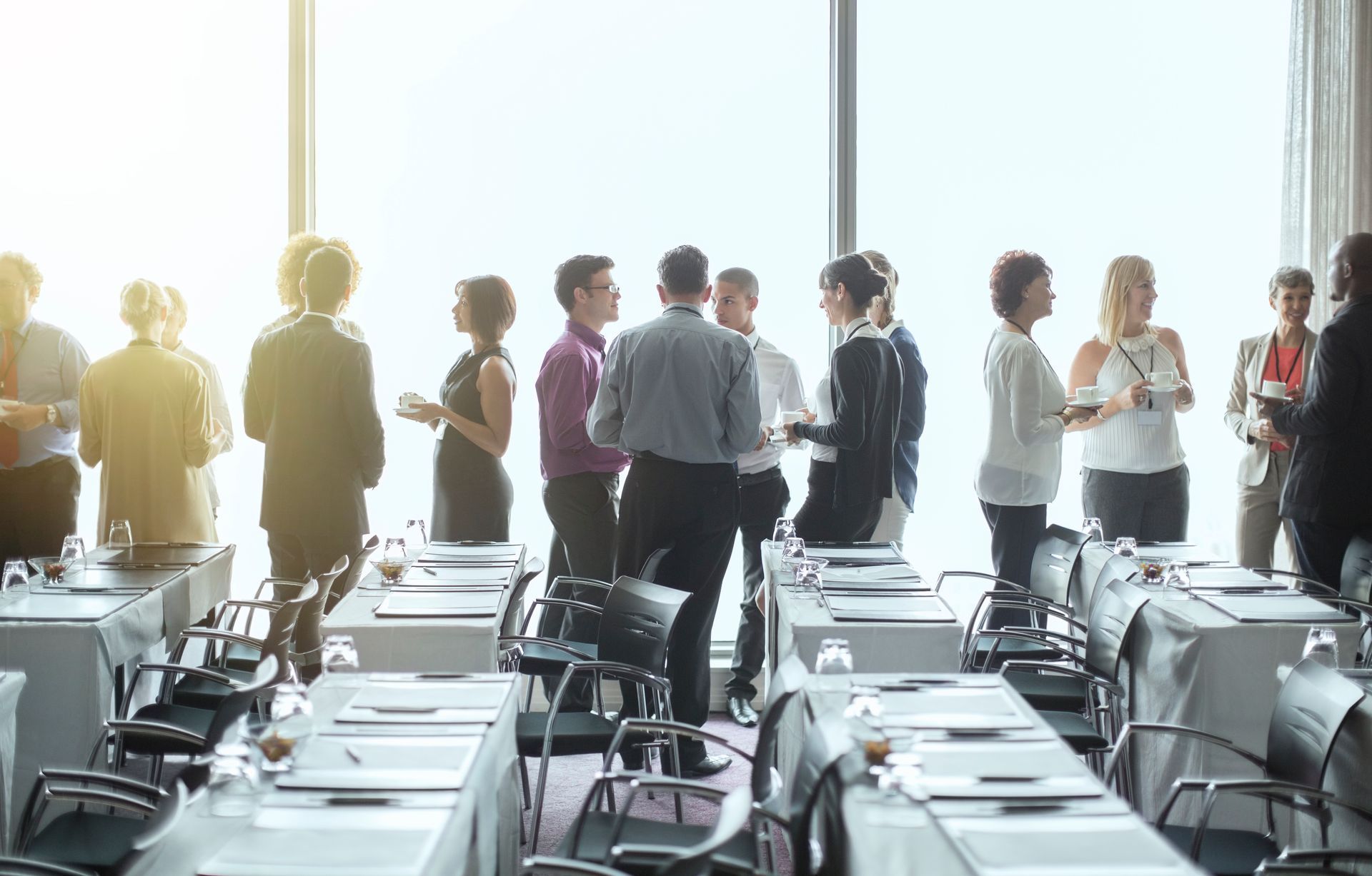 People networking at a conference, standing near tables, bright window in the background.