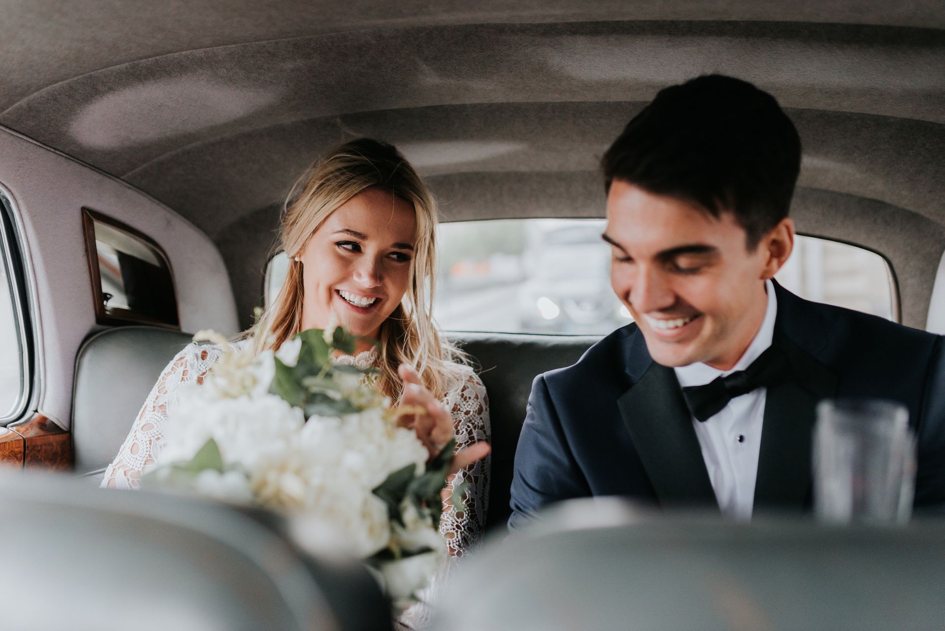 Bride and groom smile in a car; bride holds a bouquet.