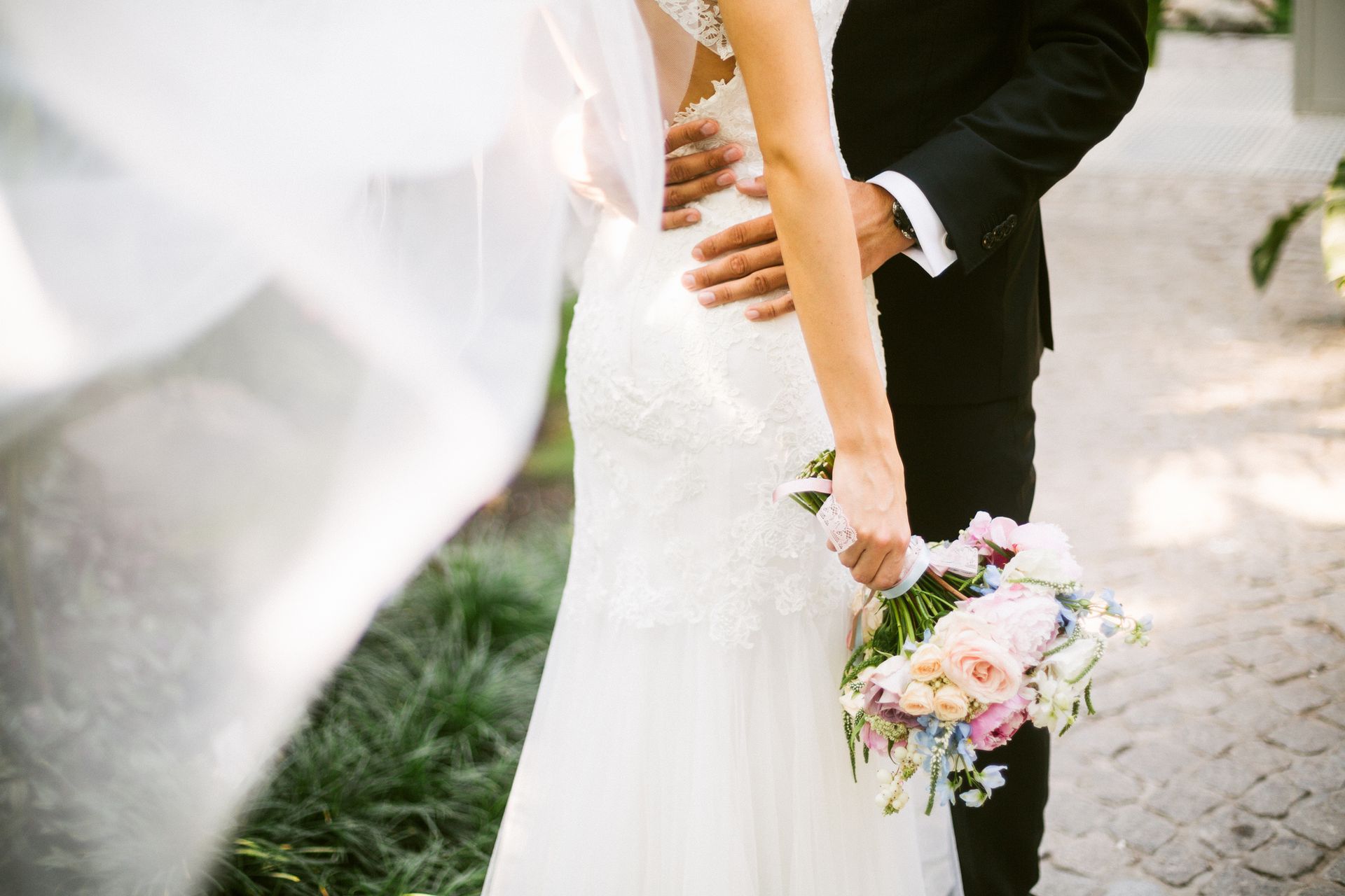 Bride and groom embrace outdoors; bride's back in white dress, groom in black suit; she holds flowers.