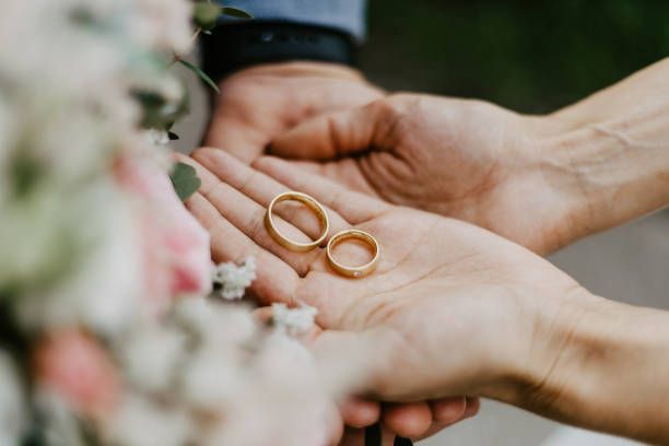 Two gold wedding rings in open palms, with a bouquet blurred in the foreground.