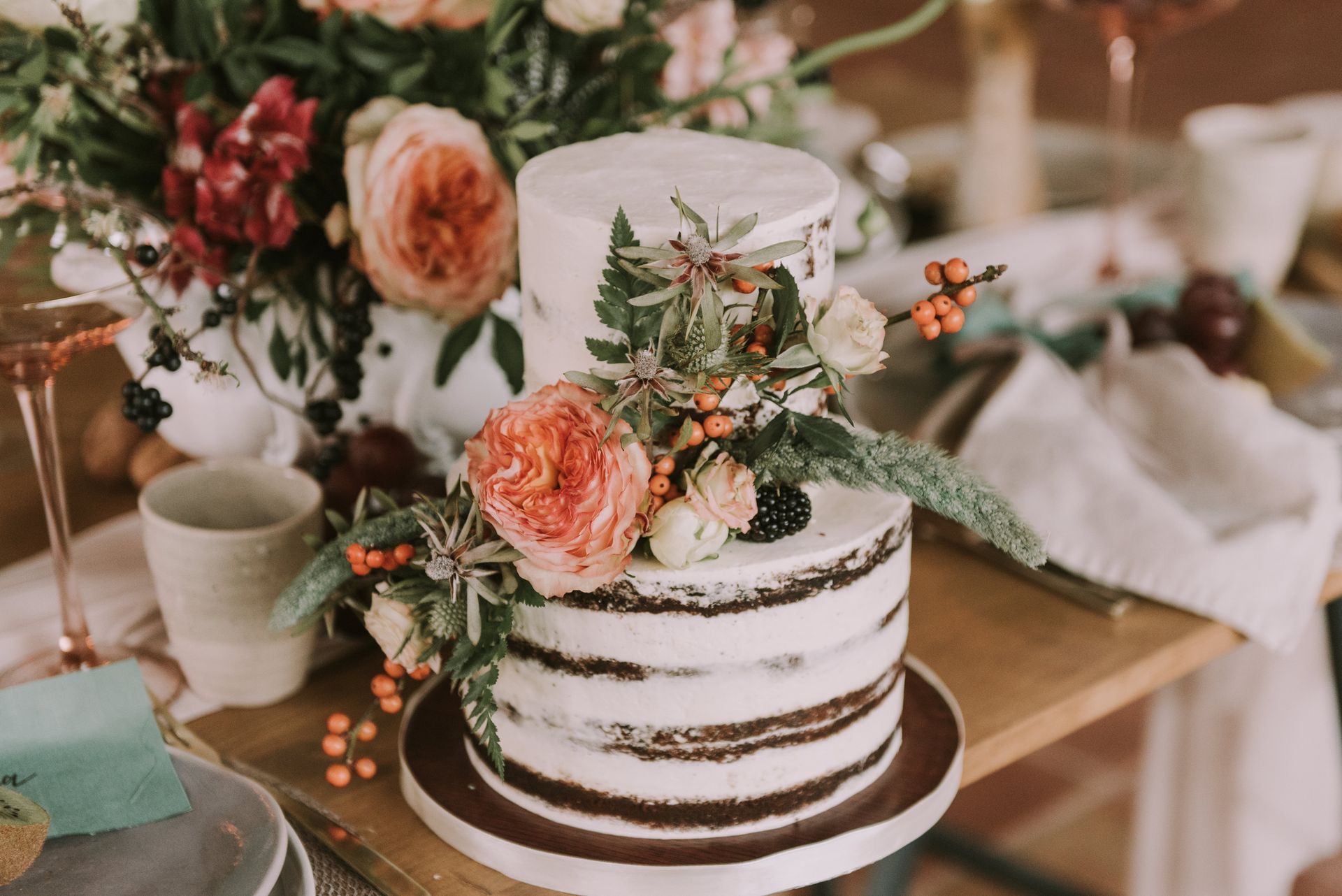 Two-tiered naked wedding cake decorated with flowers and berries on a wooden table.