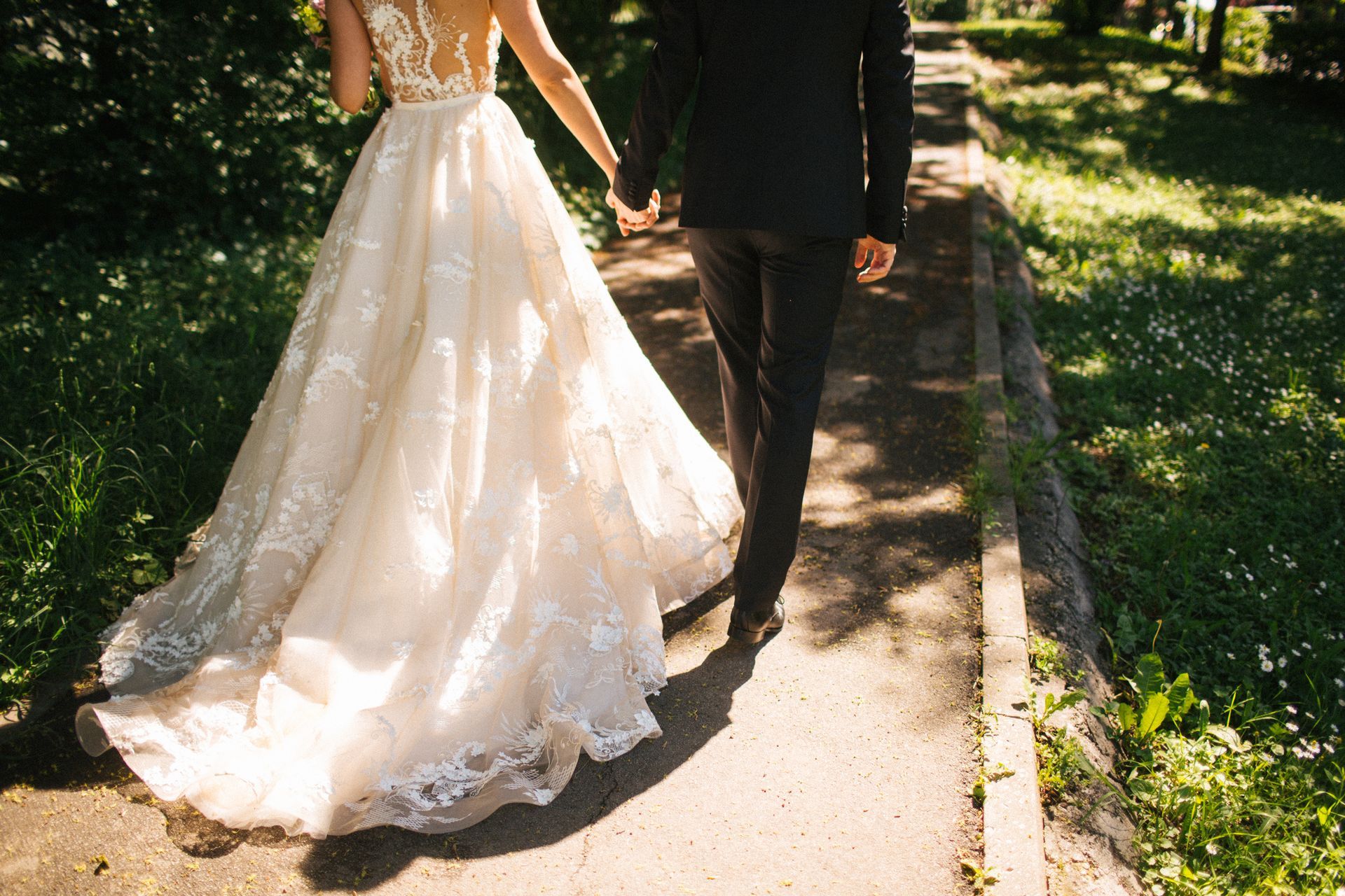 Bride in a lace wedding dress and groom in a suit walking hand-in-hand on a path in a park.