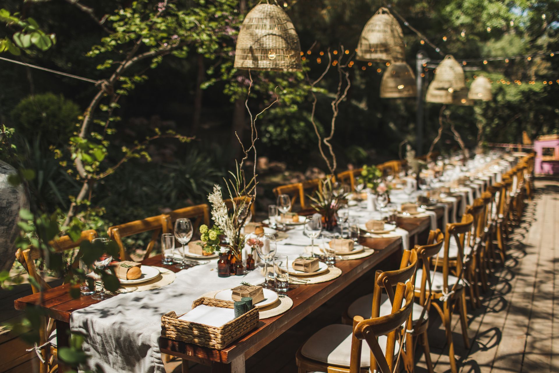 Long wooden table set for a meal in a lush garden, with decorative lighting and chairs.