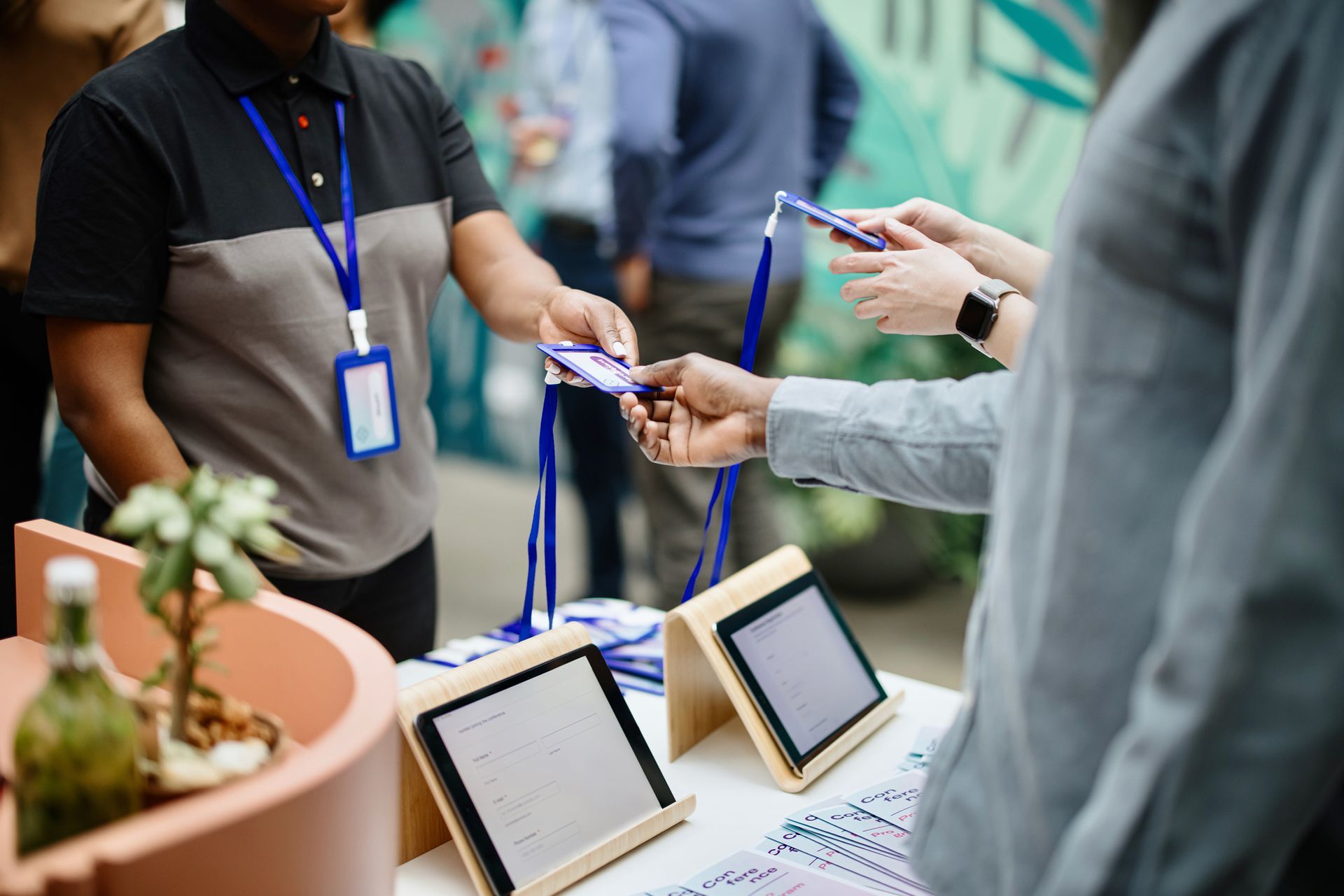 Person handing out badges at an event registration table with digital tablets and a plant.